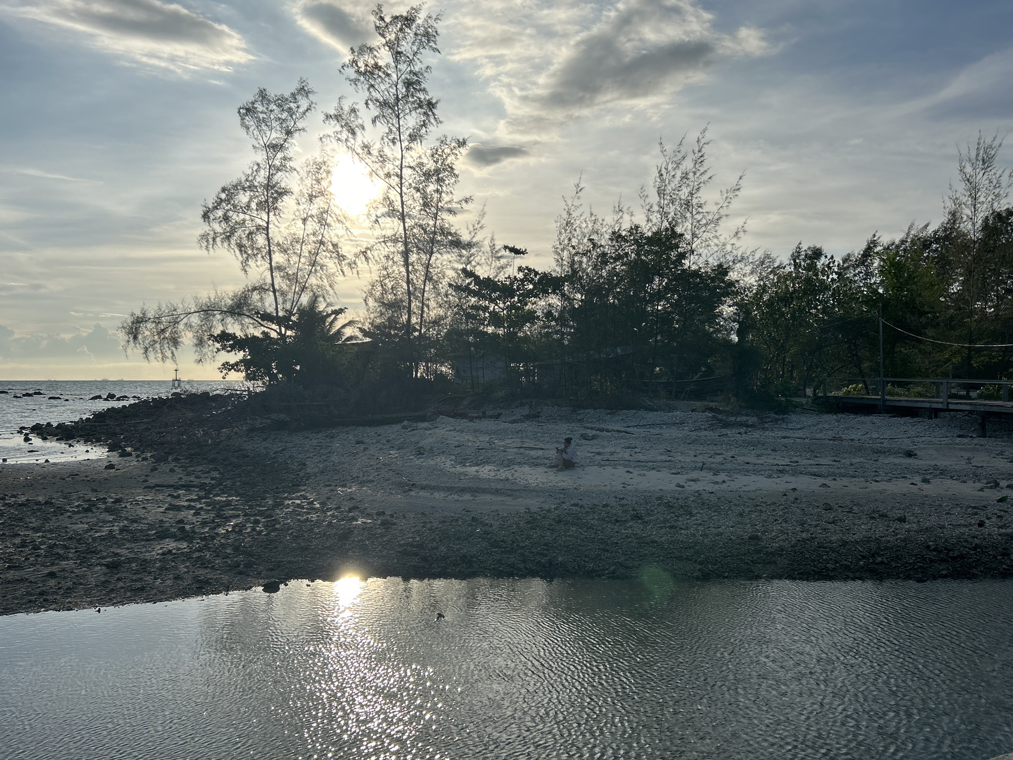 The image shows a tranquil coastal scene in ตำบลเกาะพะงัน, Thailand, at what appears to be sunset or sunrise. The sky is partially cloudy, with the sun peeking through the clouds and creating a bright glare. In the middle ground, a small island or peninsula covered in vegetation is visible. Tall, slender trees are silhouetted against the bright sky, mixed with denser patches of greenery. A person is sitting on the beach, centered and midground, but details are hard to recognize. The foreground shows a body of water, reflecting the sunlight and creating a shimmering effect. The immediate shoreline is a mix of sandy and rocky terrain. A wooden structure, possibly a pier or walkway, is faintly seen in the distance to the right. The overall scene evokes a sense of peace and natural beauty.