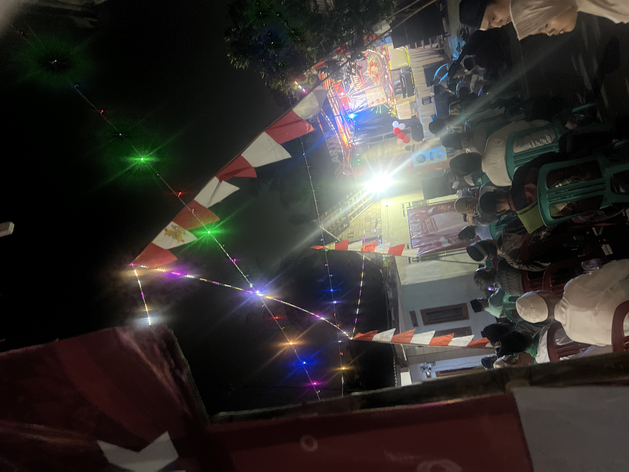The image captures an outdoor nighttime gathering in Klapanunggal, Indonesia, likely a community event or celebration. A stage is set up with a banner displaying text in what appears to be Indonesian, accompanied by colorful lights and a sound system. Red and white flags, possibly Indonesian flags, are prominently displayed, hanging from poles and strings, along with strings of multi-colored festive lights. A crowd of people, predominantly men dressed in traditional attire including peci hats and shirts, are seated in rows of chairs facing the stage. The environment appears to be a somewhat rural or semi-urban area, with simple buildings visible in the background. The lighting is predominantly from the stage and decorative lights, casting shadows and creating a festive atmosphere.