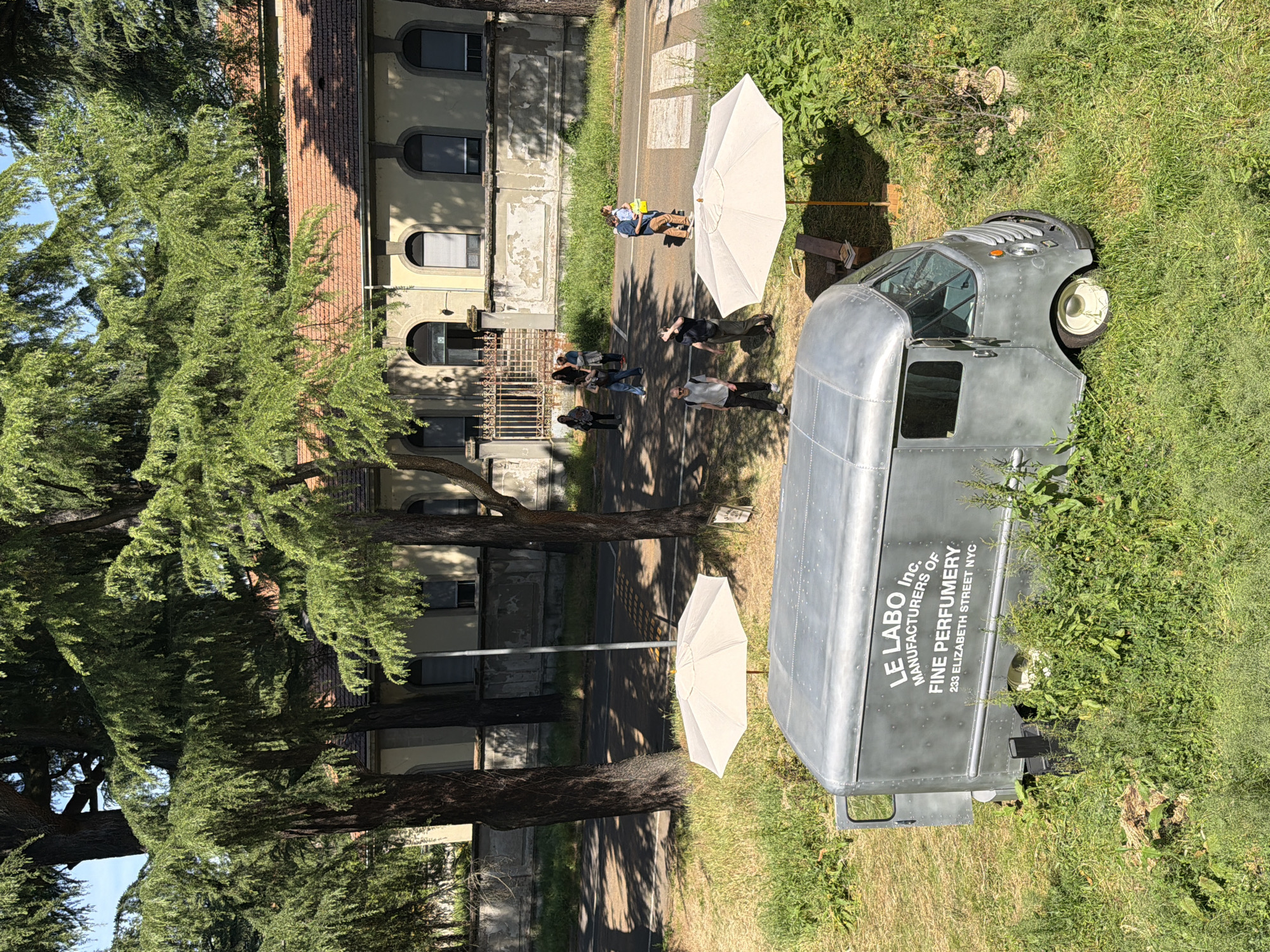 The image captures a scene in Milan, Italy, featuring a "Le Labo Inc." branded vintage delivery truck parked on a grassy verge next to a building. A group of people are standing and conversing on a paved walkway nearby. Two white umbrellas provide shade for the group and the parked truck. The building appears to be an older structure with arched windows and a weathered façade. In the background, other individuals can be seen walking along the path. The surrounding environment is characterized by trees and vegetation.