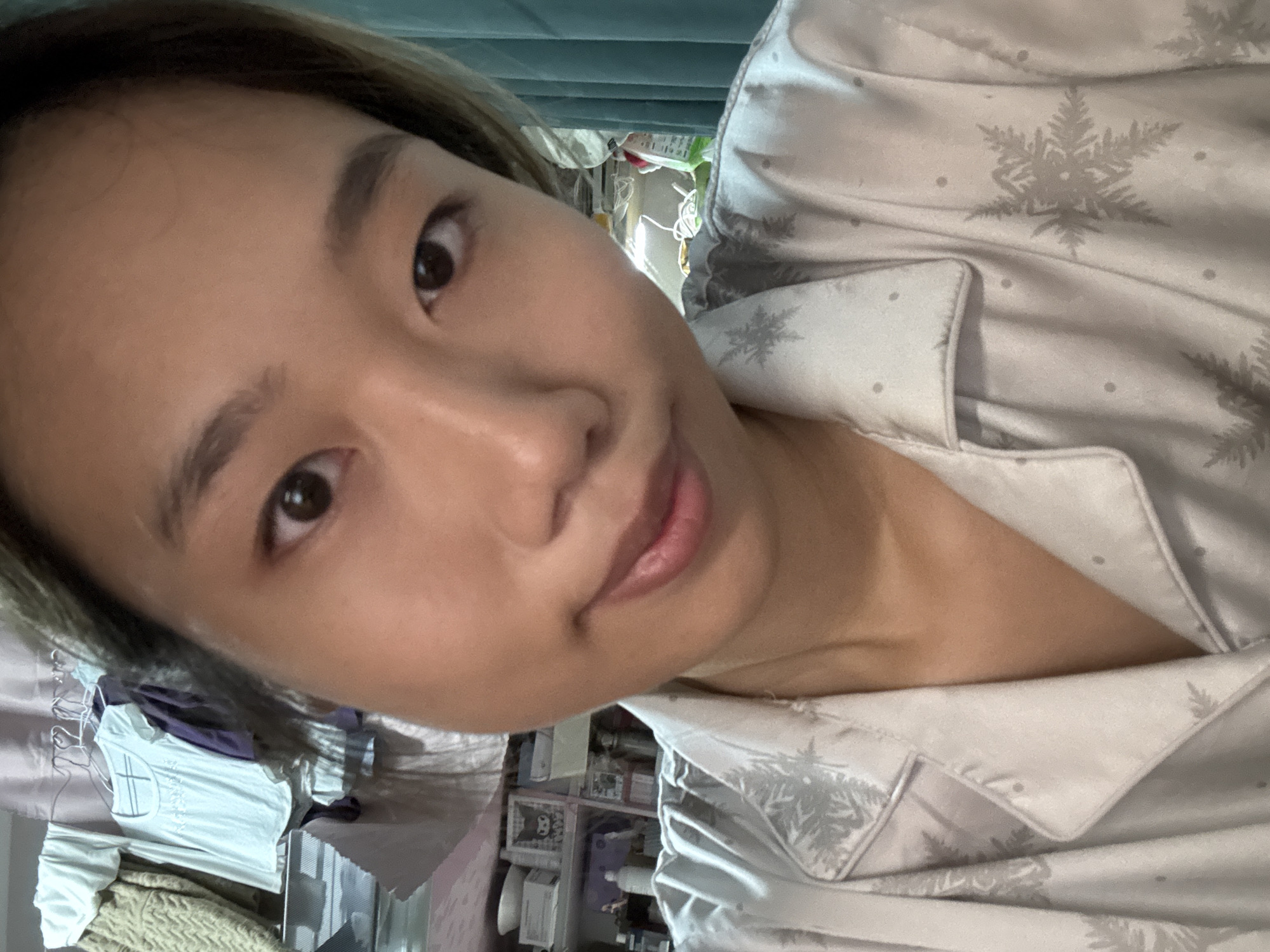 The image is a close-up, high-angle selfie of a young woman indoors. She is wearing light-colored pajamas with a snowflake pattern. Her hair is brown and appears to be pulled back, with some strands falling forward onto her face. Her eyes are dark brown, and she has a neutral expression. The background is slightly out of focus but shows what appears to be a cluttered room with shelves filled with various items, including what looks like boxes and clothing. The lighting suggests it is daytime. The overall mood is casual and relaxed, possibly taken in a bedroom. There is no visible text in the image.
