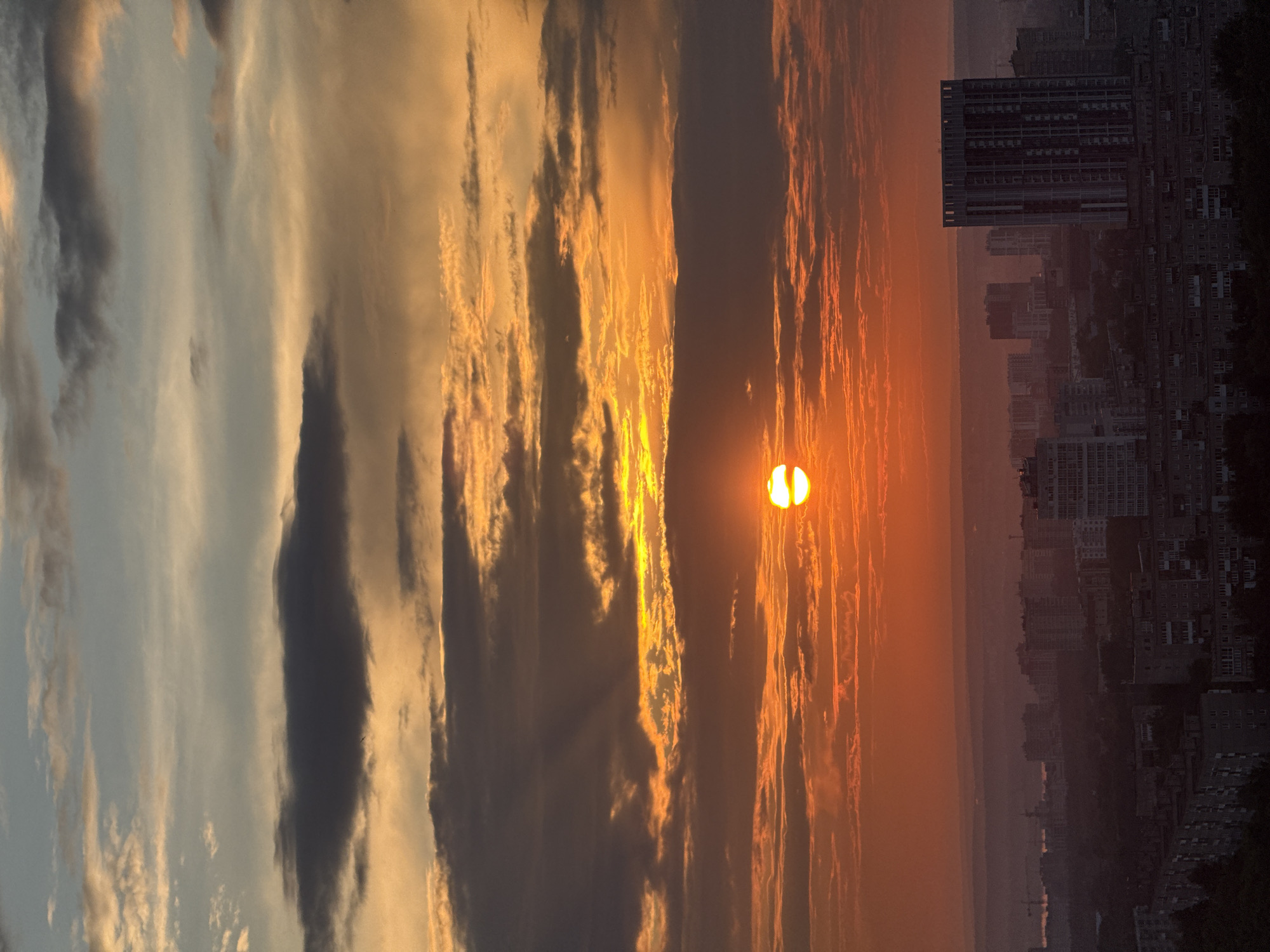 The image depicts a sunset over the city of Zarechnaya (Gamovskoe S/P), Russia. The sky is filled with dramatic clouds in shades of orange, yellow, and gray, with visible sun rays piercing through. The sun, a bright orange orb, is setting near the horizon. Below the sky, the cityscape is visible, characterized by numerous buildings of varying heights and architectural styles. A prominent tall, modern building with many windows stands out among the others. The foreground shows a dense collection of lower-rise residential buildings, casting shadows. The overall impression is a serene, urban landscape bathed in the warm, diffused light of the setting sun.