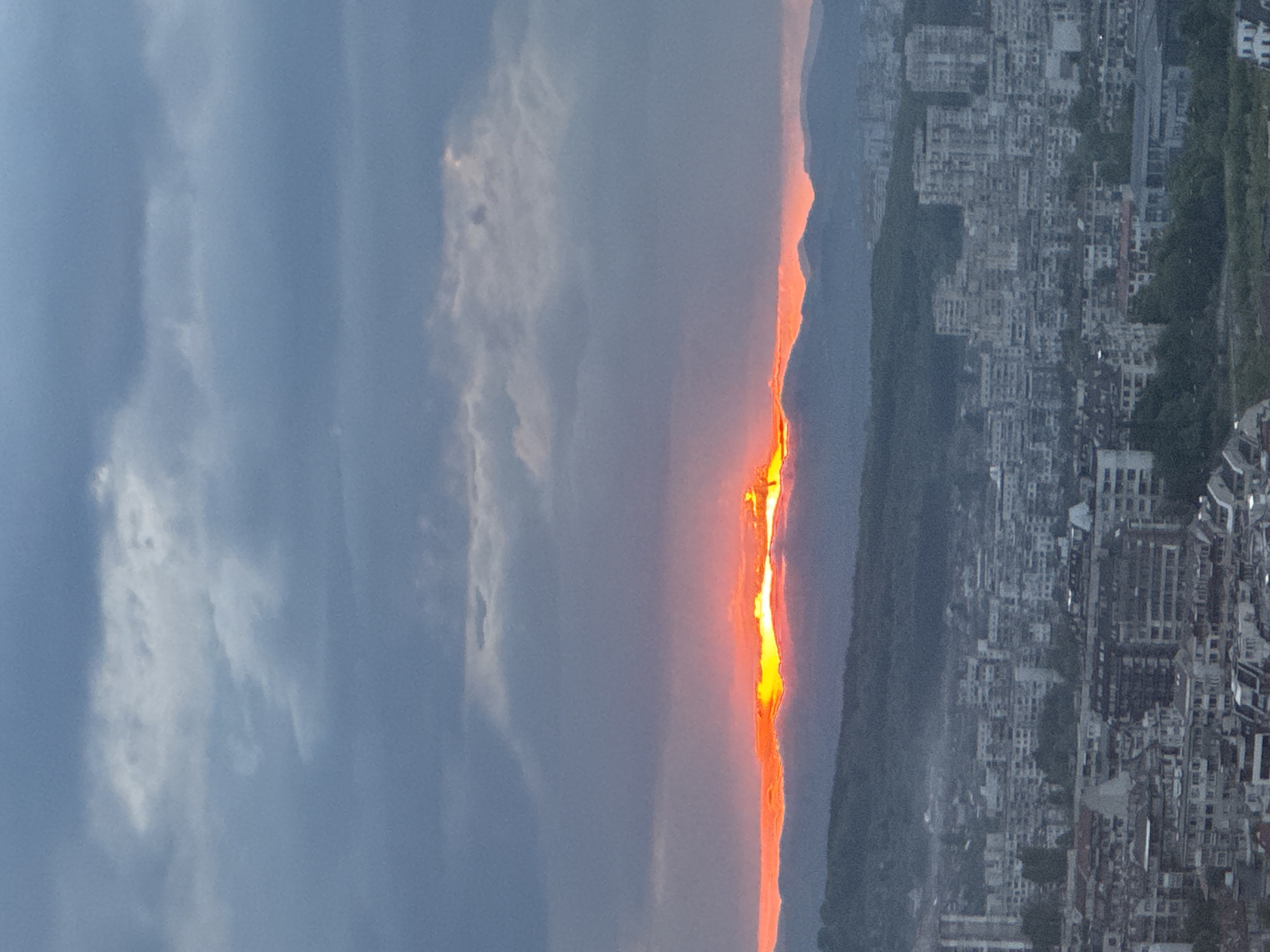 A vertical-oriented, high-angle aerial shot depicts a cityscape at sunset, likely in Sofia, Bulgaria, viewed from a high vantage point. To the left, the sky is filled with blue and grey clouds. A vertical band of orange and yellow sunlight pierces through the clouds near the center of the image, highlighting the edges of mountains or hills in the distance. To the right, the cityscape is visible, filled with numerous buildings that vary in size and density, many of which appear residential. Some green vegetation is visible among the buildings. The setting appears urban and partially hilly.