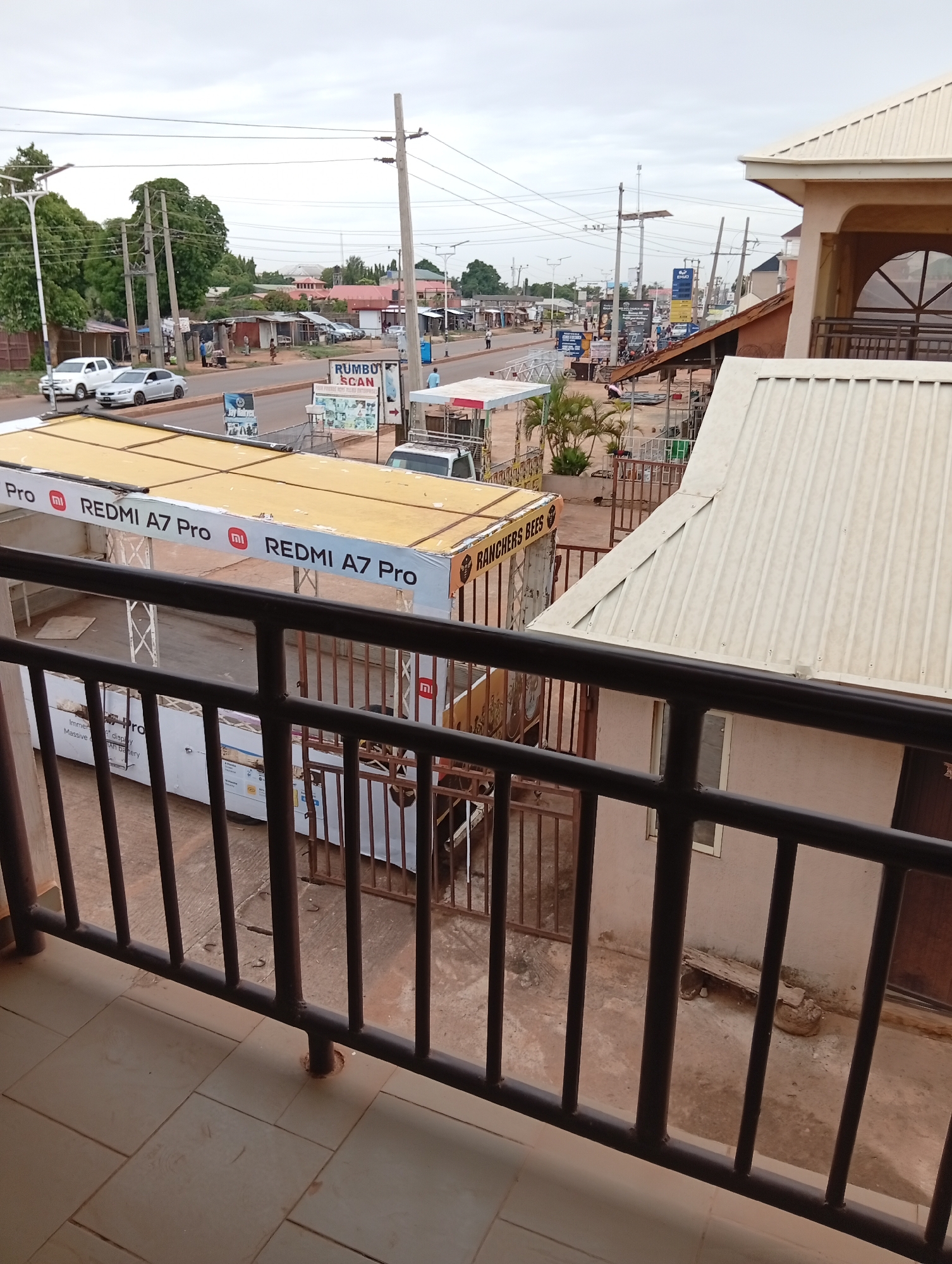 The photograph depicts a street scene in Sabon Tasha, Nigeria, viewed from an elevated position, likely a balcony. A metal railing is in the foreground. Visible beyond the railing is a street with vehicular traffic, including cars and trucks. Commercial establishments line the street, including vendors' kiosks with "REDMI A7 Pro" and "RANCHERS BEES" signage. Utility poles and power lines stretch along the street. People can be seen walking and standing on the sidewalks. The buildings on the right side of the frame have a two-story structure, including an arched window detail. The sky is overcast.