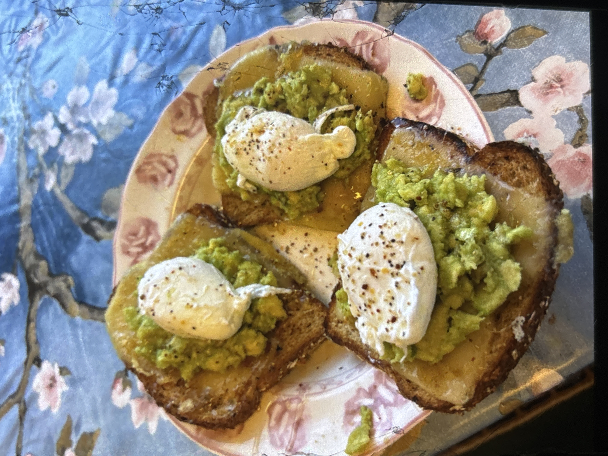 Three pieces of toast topped with mashed avocado and a poached egg are presented on a floral-patterned plate. The toast appears to be lightly browned and spread with a green, textured avocado mixture. Each piece of toast is crowned with a single, oval-shaped poached egg, seasoned with black pepper. The plate sits on a table with a blue and pink floral tablecloth. The image is taken from a slightly elevated angle, capturing the texture of the food and the details of the plate and tablecloth. There are no people visible in the image. The location is not specified in the image itself or the provided context, so any mention of Orihuela, Spain would be an assumption.