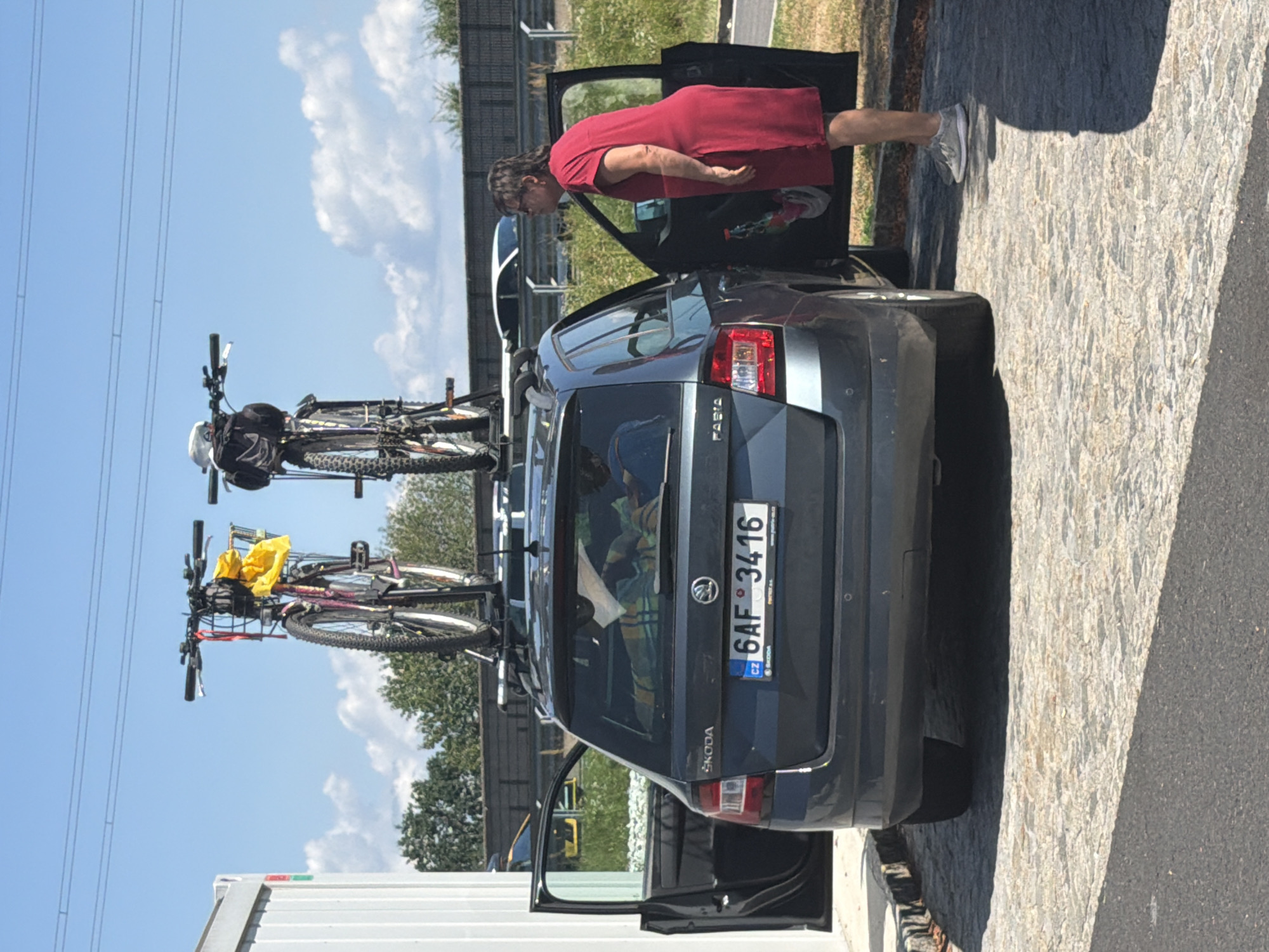 An outdoor, eye-level medium shot shows a dark gray Skoda Fabia station wagon parked on a cobblestone surface. The car's rear is visible, with the driver's side door open. A woman wearing a long, red dress and gray sneakers stands beside the open car door, facing away from the camera. Two bicycles are mounted on a roof rack atop the station wagon. The sky is blue with scattered white clouds. In the background, a white barrier and trees are visible. A license plate on the rear of the car reads "6AF 3416". The image is captured during daylight. The exact location is not identifiable within the city of Gralla, Austria, based on the visual information provided.