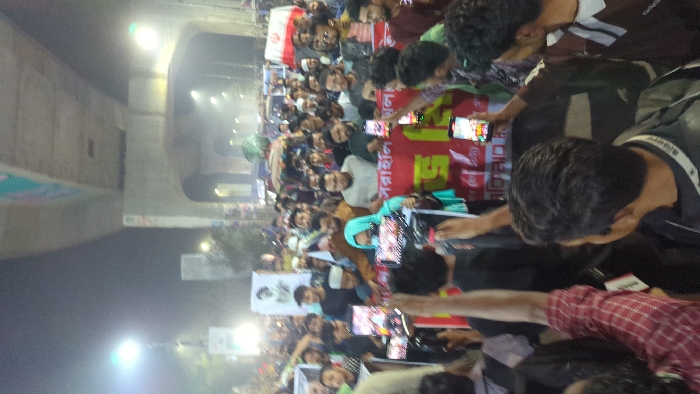 The image captures a dense nocturnal gathering of people on a street in Dhaka, Bangladesh, beneath an elevated concrete flyover or metro rail structure. The scene is illuminated by multiple bright artificial light sources, casting a slight haze.
The crowd is predominantly composed of young to middle-aged males, packed closely together and extending deep into the background. Several individuals wear skullcaps or traditional attire. A female wearing a turquoise hijab is visible in the mid-ground. Numerous participants are holding smartphones aloft, actively recording or photographing the event.
A large red banner with yellow Bengali script is stretched horizontally across the mid-ground; visible text includes "সরাহনি" (condemnation/protest) and "আহ্বান" (call/appeal). In the upper-left mid-ground, a white sign displays a black-and-white portrait of a man. A white flag with a red emblem is also visible among the crowd. The concrete pillars supporting the overhead structure are prominent in the upper frame. The gathering appears to be a public rally or protest.