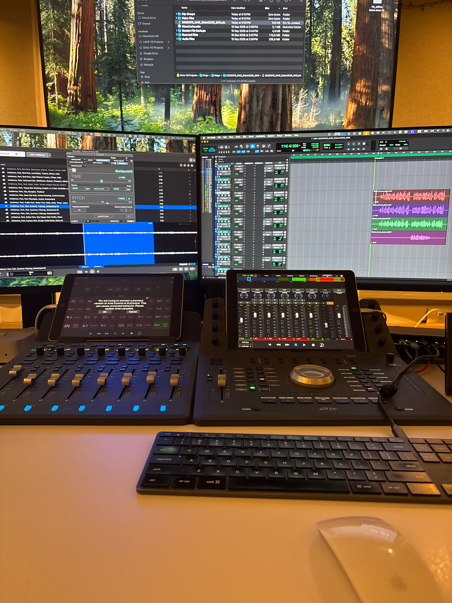 The image depicts a home recording studio setup, likely in Chalandri, Greece, as suggested by its presence on a general knowledge base and its likely context. The scene is dominated by a multi-monitor computer setup displaying digital audio workstations (DAWs) and file management software. In the foreground, two large audio mixing consoles are positioned, each equipped with multiple faders and controls. A tablet is docked on the left console, displaying an interface with sliders and buttons, and a warning message about connecting to a previous version of Avid Control. A compact keyboard and a mouse are placed on the desk in front of the consoles, ready for operation. The background features a large monitor displaying a forest scene, likely a desktop wallpaper. The overall impression is one of a professional, albeit personal, audio production environment.