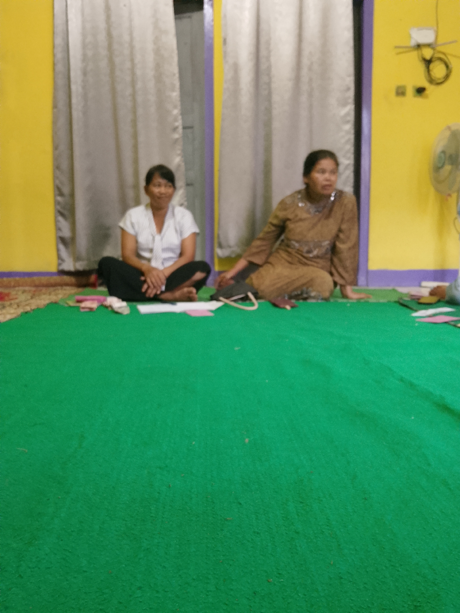 Two women are seated cross-legged on a vibrant green carpet in what appears to be a room with yellow walls and light grey curtains. The woman on the left wears a white collared shirt with thin pinstripes and black pants. Her hands are clasped in her lap. The woman on the right is dressed in a brown patterned tunic and is looking to her right. A fan is visible on the right side of the frame. There are some papers and what look like folded banknotes on the carpet in front of the seated women. The overall lighting is somewhat dim. The scene is set in Jumo, Indonesia.