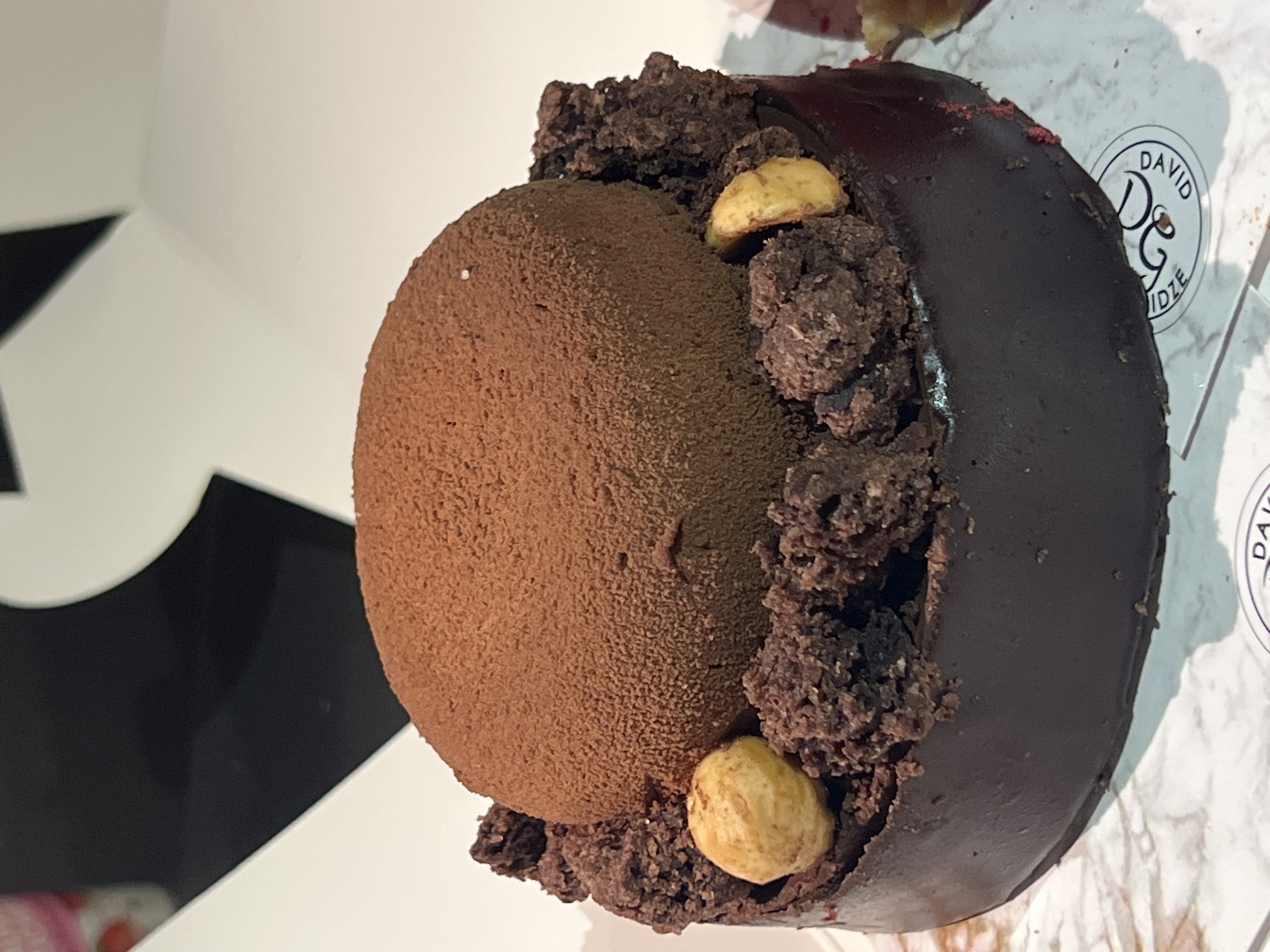The image displays a close-up, slightly tilted view of a decadent chocolate dessert. The primary subject is a round pastry or cake, featuring a prominent spherical top generously dusted with cocoa powder, creating a matte, velvety brown texture. The lower portion of the dessert is encased in a smooth, dark, and subtly glossy chocolate glaze. Around the base of the cocoa-dusted dome, there are scattered pieces of dark chocolate crumble, resembling soil, which add a contrasting texture. Two whole, light-colored nuts, likely hazelnuts, are embedded within these chocolate crumbles.
The dessert rests on a sheet of white paper with a subtle marbling pattern, likely part of its packaging. In the background, elements of a white box with stark black geometric shapes are partially visible. On the white paper next to the dessert, a circular logo is clearly visible. The logo contains the text "DAVID D&G IDZE", with "DAVID" positioned at the top, "IDZE" at the bottom, and a stylized "D&G" in the center. The scene captures a still life of this confectionery item, presented in what appears to be takeaway packaging, suggesting it's from a bakery or patisserie in Tbilisi, Georgia.