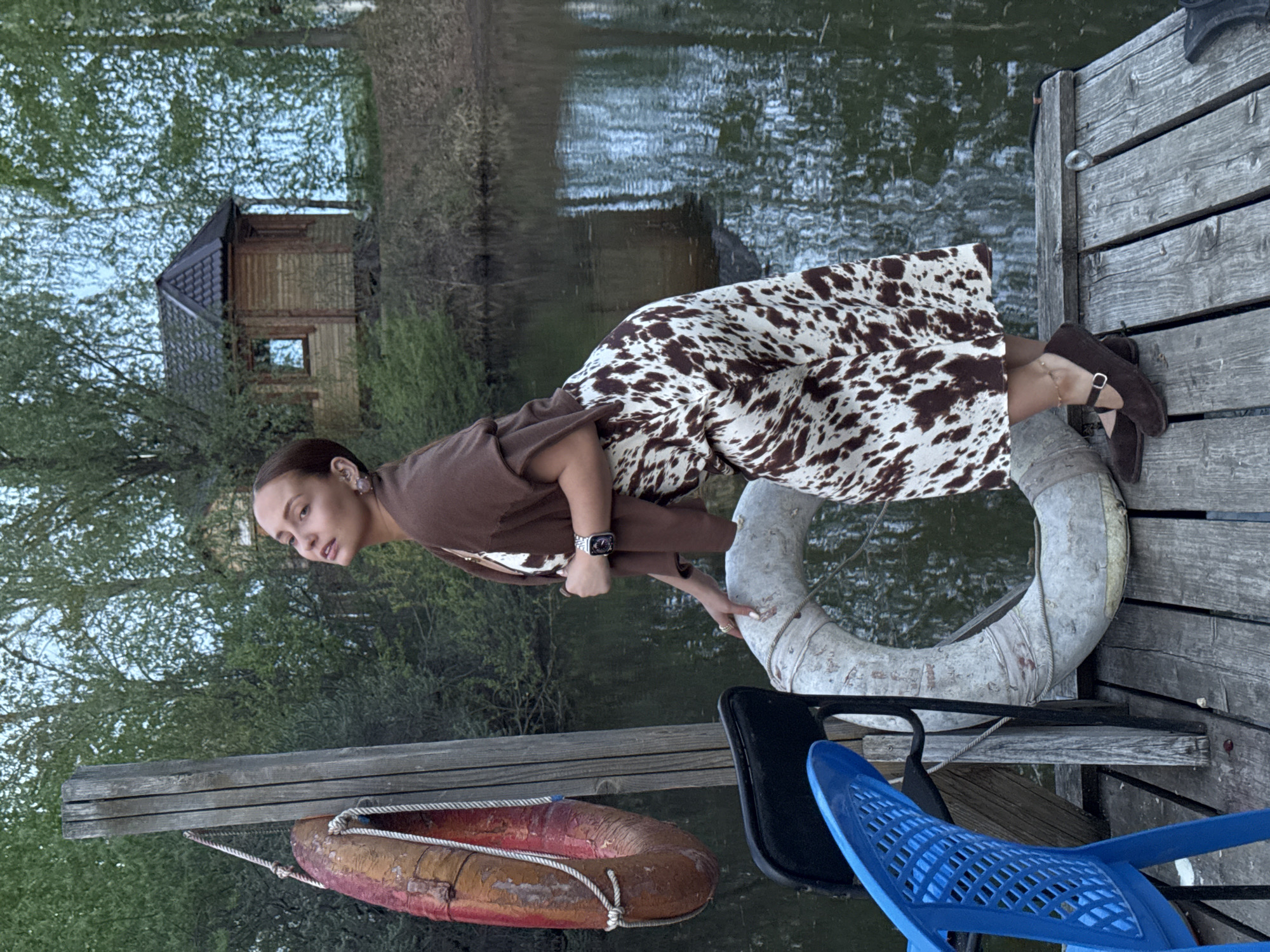 A young woman is on a wooden dock, wearing a brown dress with a cow-print pattern and brown shoes. She is leaning forward, holding onto a white life preserver. Below her and to the left is a red and orange life preserver hanging from the dock. Nearby are black and blue plastic chairs. In the background, there is a body of water with a small wooden structure on the opposite shore. The location is Chaikovsky, Russia.