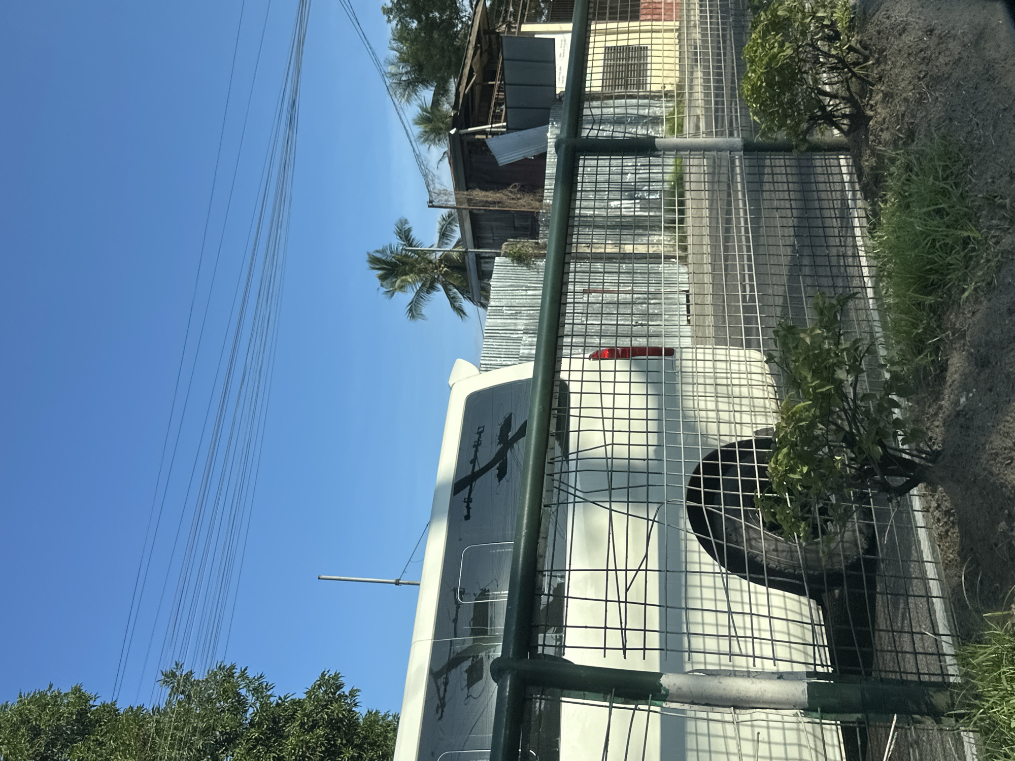 This image captures a bright daytime scene in Davao City, Philippines. A dark green metal mesh fence with green and white striped posts occupies the lower and mid-ground, separating the viewer's immediate space from the activity beyond. In the mid-ground, a white van is partially visible behind the fence on the left, displaying its side profile with dark tinted windows. These windows reflect intricate patterns of overhead utility poles and wires, distinct from the power lines in the sky. The van's rear passenger wheel is visible, and a sliver of a red taillight is discernible on the far left.
Beyond the van and the primary fence, a second, corrugated metallic fence runs horizontally, bordering a street or paved area. Behind this second fence, various structures are present. On the left, a building constructed from weathered wood and corrugated metal is partially obscured by lush green foliage and several palm trees. To the right, a cream-colored building with a dark rectangular window is visible. The upper half of the image features a clear, bright blue sky, intersected by multiple parallel black power lines. A slender silver pole extends vertically into the sky from the lower-middle left. The foreground consists of dirt patches with sparse green grass and small cultivated bushes near the fence posts. No people are visible.
