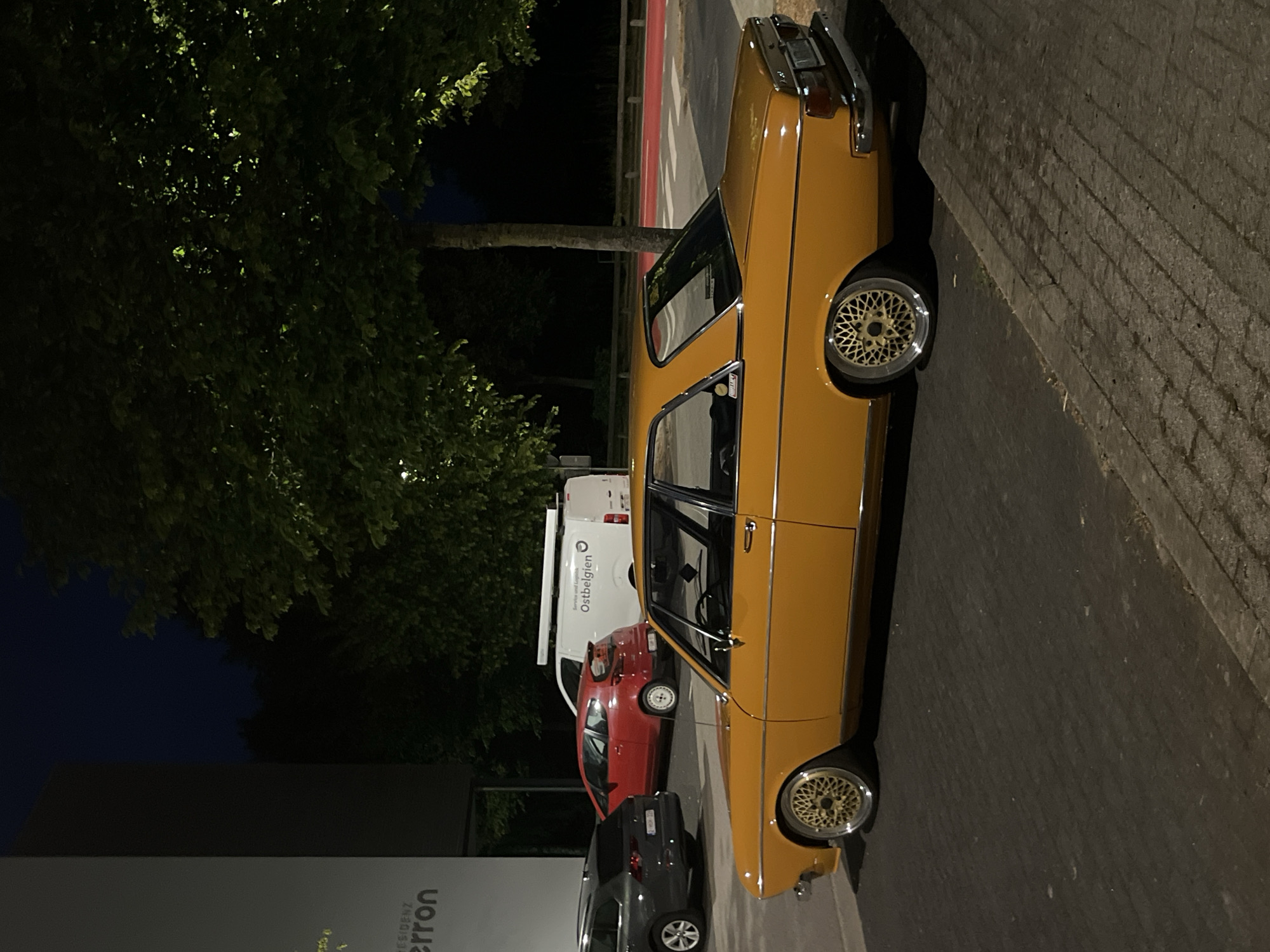 The image depicts an outdoor scene at night in the city of Saint Vith, Belgium. The primary subject is a vintage, bright yellow sedan parked on an asphalt surface. The car features chrome accents and distinctive gold wheels with a mesh design. To the left of the sedan, a dark gray SUV is partially visible. Further in the background, a red hatchback and a white commercial van are parked. The van has text on its side that reads "Service und Logistik Ostbelgien".
The environment includes a building with a dark facade on the far left, with some text visible that includes "erron". Several trees with dense foliage are present, with some leaves illuminated by external light sources. The background reveals a dark sky, suggesting nighttime, and a red-lined pathway or road. The ground surface is a mix of asphalt and brick paving stones. No people are visible in the image, and no actions or interactions are occurring. The scene appears to be a street or parking area.