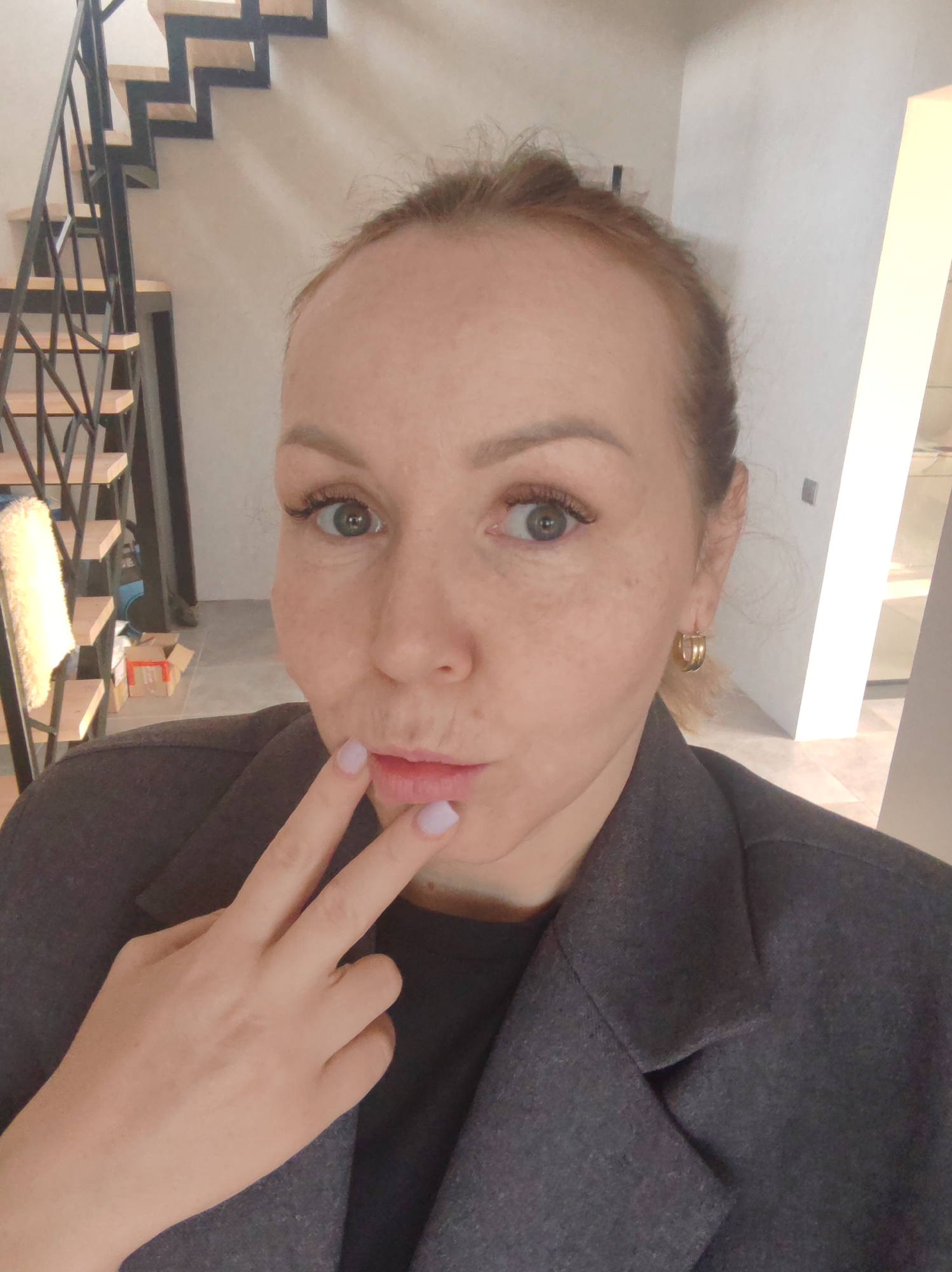 A person with fair skin, light red hair pulled back, and wearing a grey blazer and gold hoop earrings is taking a selfie. The person is holding two fingers up to their lips, which are slightly puckered. Freckles are visible on their face.
Behind the person is an interior view of a building with a modern staircase featuring black metal railings and wooden steps. A white wall is behind the staircase. A light-colored door or entryway is to the right.
There are no indications of the city of Chaikovsky, Russia, in the image.