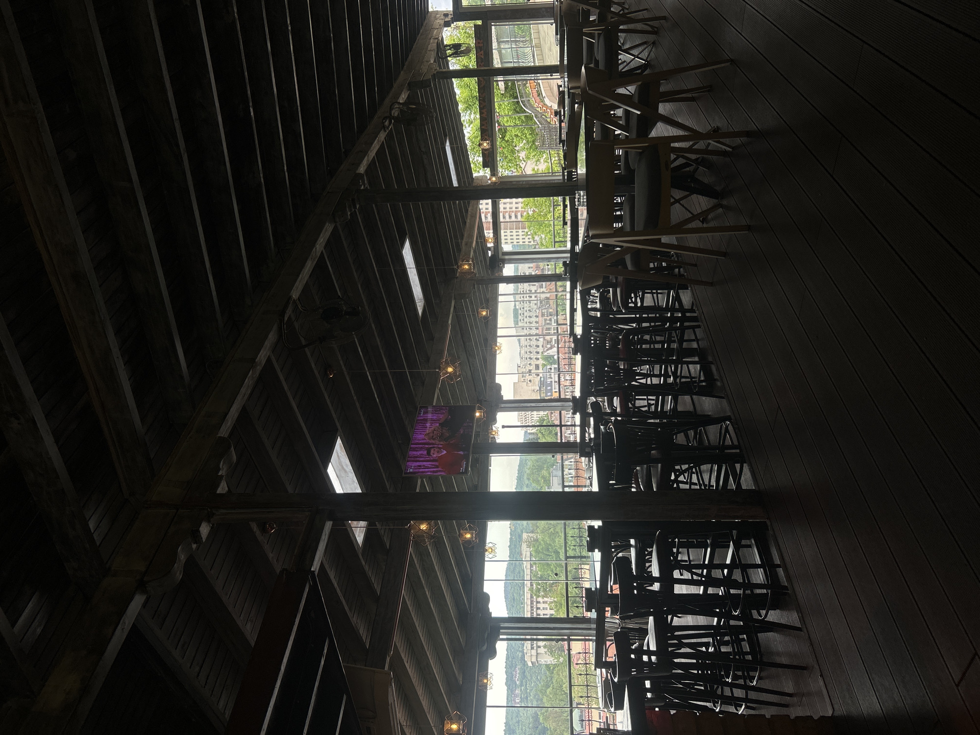 The image shows the interior of a restaurant with wooden floors and ceiling beams. Rows of tables and chairs are arranged along the windows. A television screen is visible on one of the supporting beams. Outside the windows, the cityscape of Veliko Tarnovo, Bulgaria is visible. No people are clearly visible.