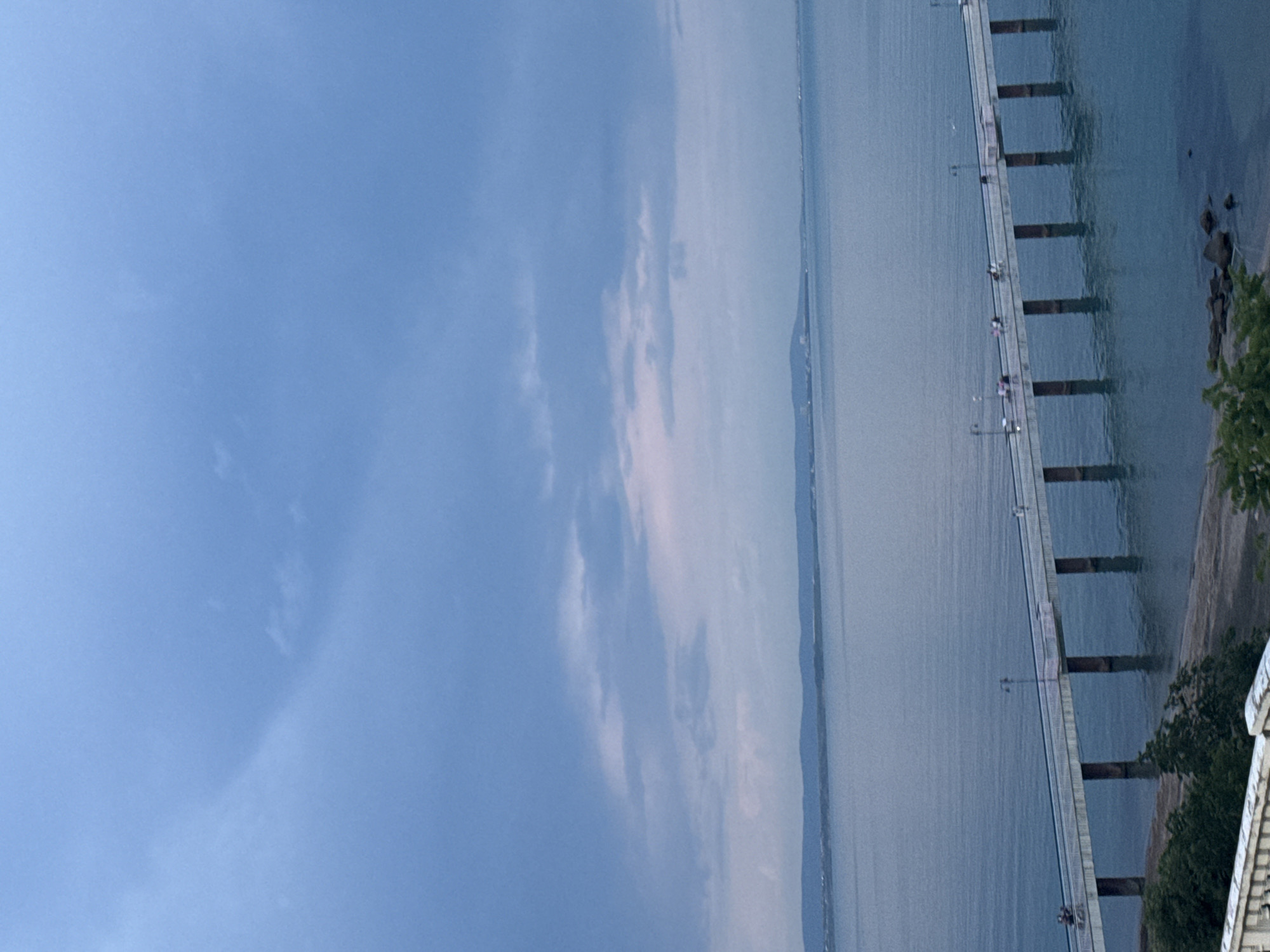 The image shows a pier stretching out into the water, likely in Burgas, Bulgaria. The pier is constructed of concrete pillars supporting a flat walkway. Several people are visible walking and standing along the pier. The water is calm, with a hazy line of land visible in the distance. The sky is predominantly blue with patches of cloud cover. Vegetation is present on the shoreline near the pier.