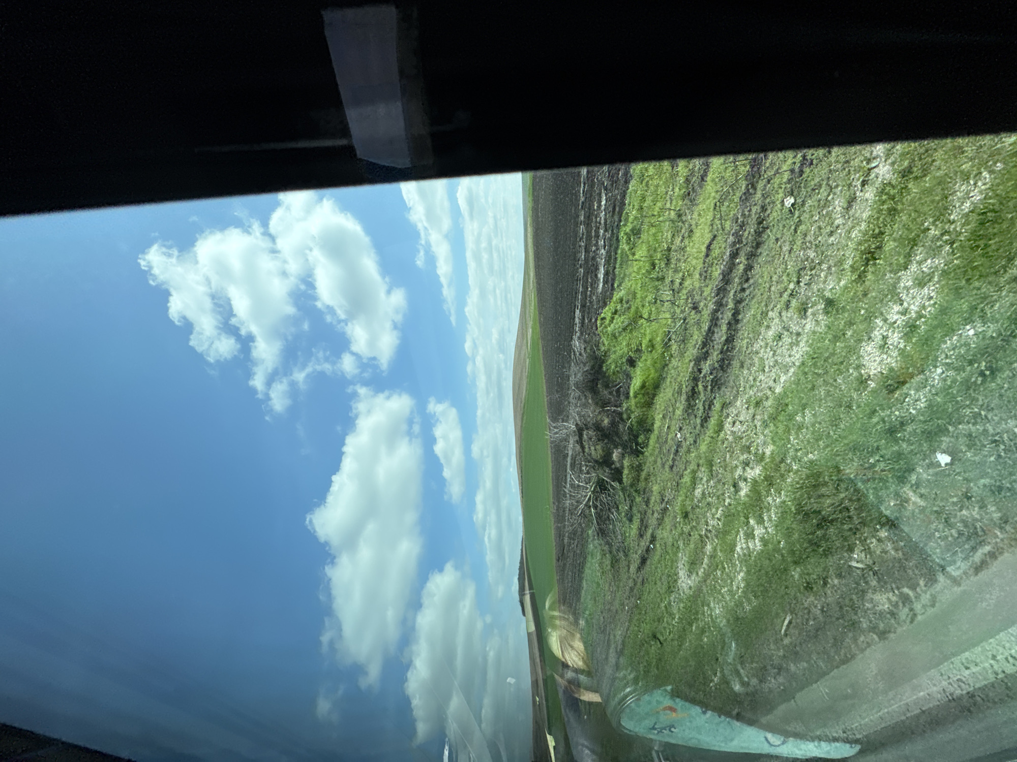 The image shows a view from inside a vehicle, likely a car or truck, looking out through the windshield. The sky is blue with scattered white clouds. The landscape features a mix of terrain including cultivated fields (some with green crops, others recently plowed and dark brown), and grassy areas. The vehicle's interior is visible at the top and bottom of the frame. The location is unspecified beyond a view of outdoor terrain and a sky, implying a location near Karaisen, Bulgaria. No people are visible.