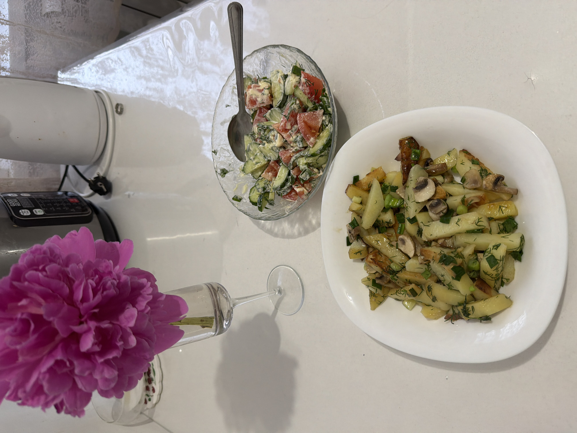 A still life is captured in a high-angle shot on a light-colored countertop. A vibrant pink peony in a glass vase is positioned on the left. A clear glass bowl holds a salad of cucumbers, tomatoes, and herbs with a spoon placed within. To the right, a white plate presents a dish of sautéed potatoes and mushrooms, garnished with dill and scallions. The countertop's surface has a subtle textured pattern. A kitchen appliance is partially visible in the upper-left corner. The image was taken in Leningradskaya, Russia.