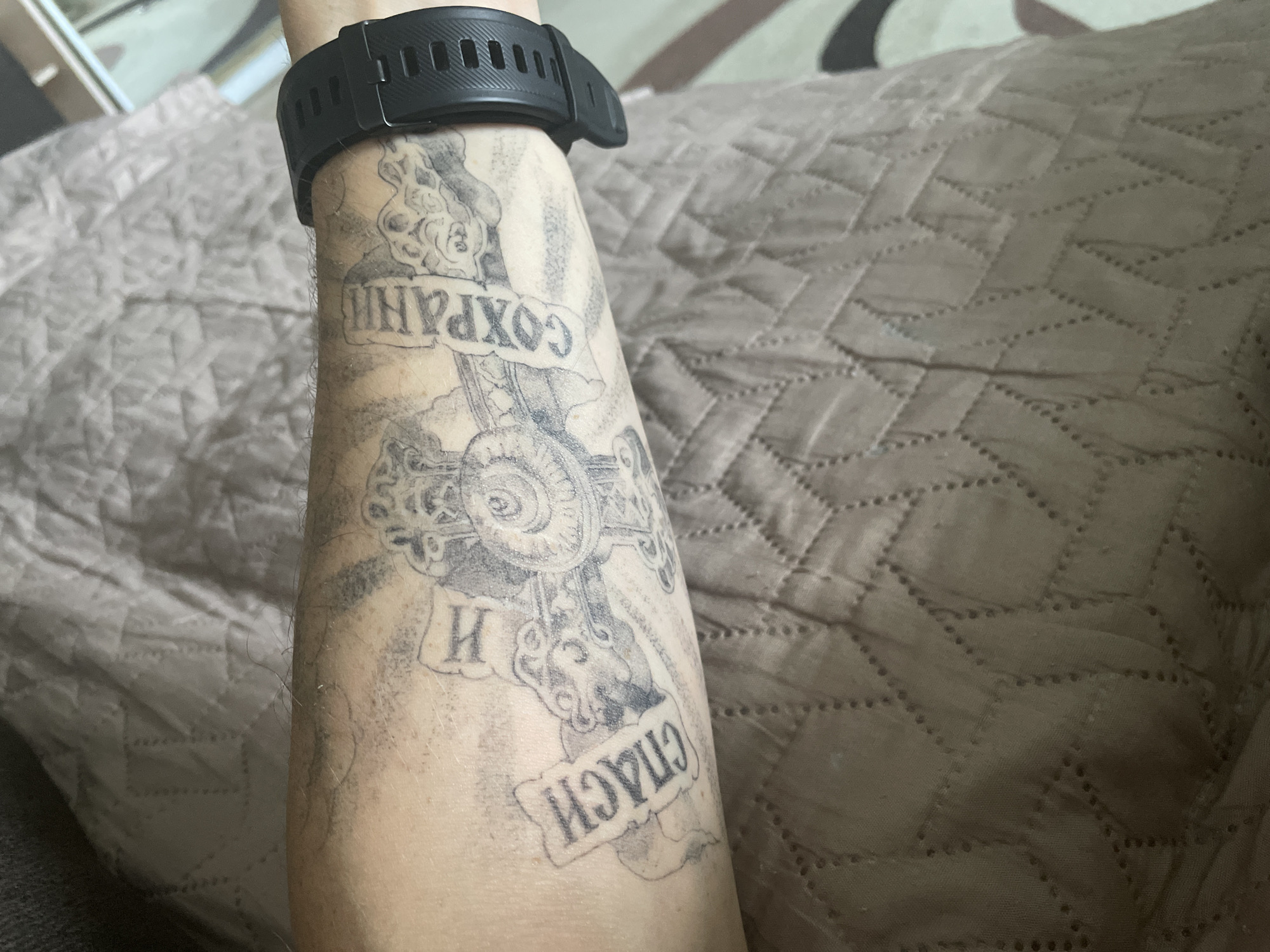An arm with a tattoo of a cross and Russian text is visible against a quilted fabric background. The tattoo is black and gray and appears to read "СПАСИ И СОХРАНИ" (Save and Protect) with the letters "И" and a partial "С" visible. A black smartwatch is worn on the wrist above the tattoo. The arm is positioned on a brown quilted blanket. There is no visible indication of the location being in Sarapul, Russia.