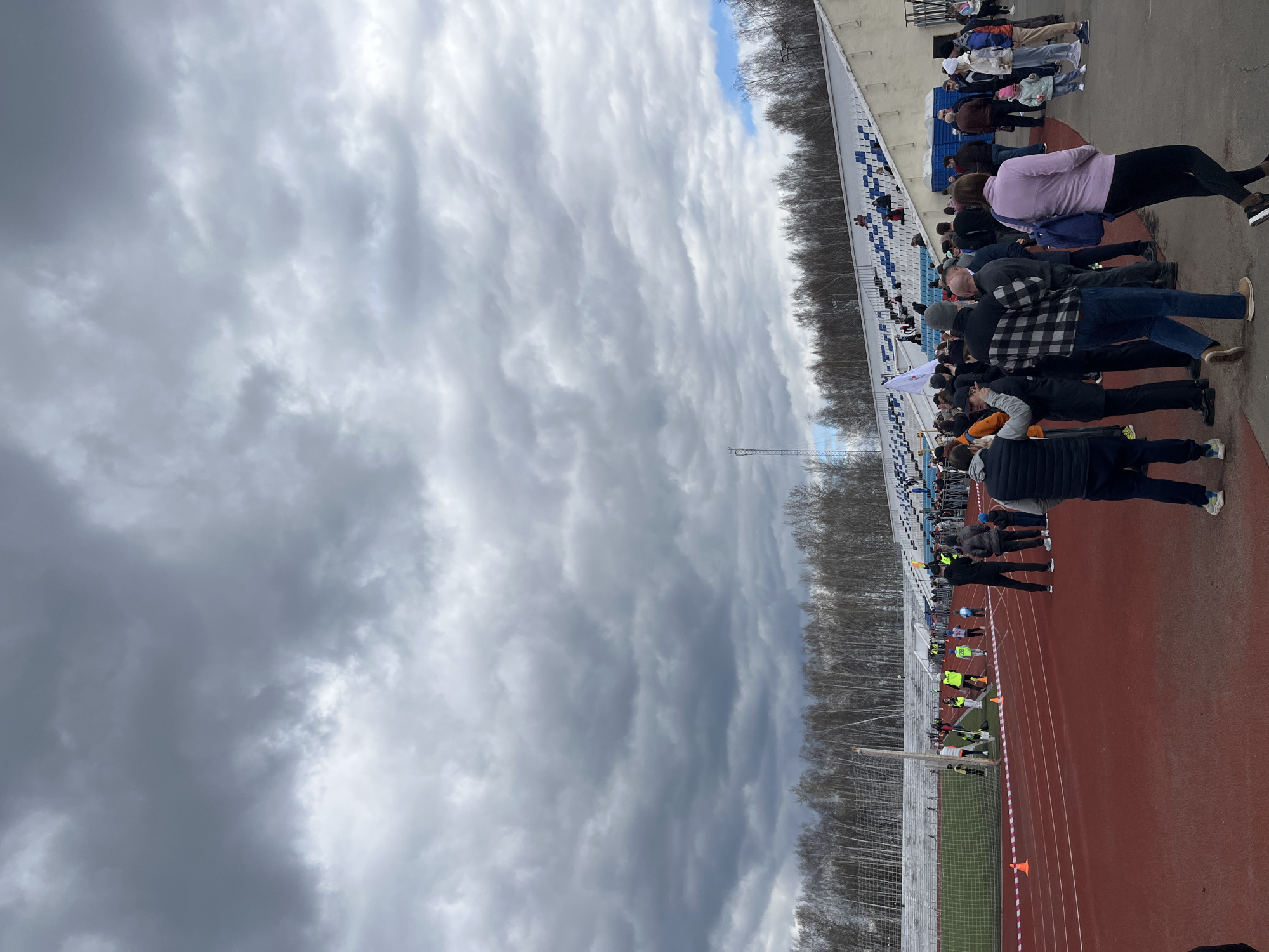 The image shows a view of a stadium in Chaikovsky, Russia, under a cloudy sky. There is a running track in the foreground with athletes and officials gathered near the start line. Spectators are present in the stands overlooking the track. Trees line the perimeter of the stadium behind the stands.