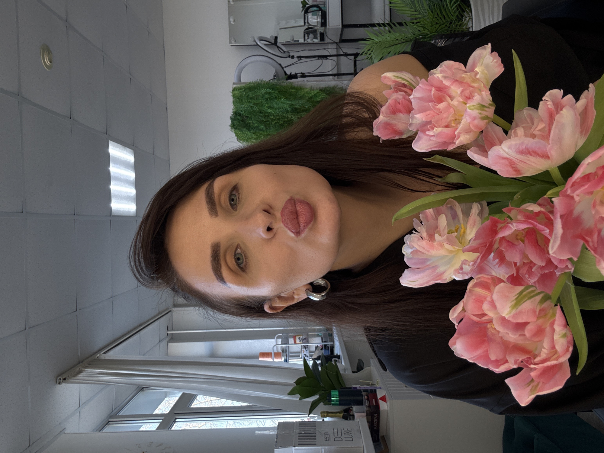 A woman with brown hair and pink lipstick is holding a bouquet of pink tulips in an indoor setting. The location appears to be a room with tiled walls, a window with blinds, and various items on a shelf, including cosmetics and plants. The city of Chaikovsky, Russia, is not identifiable from the image alone.