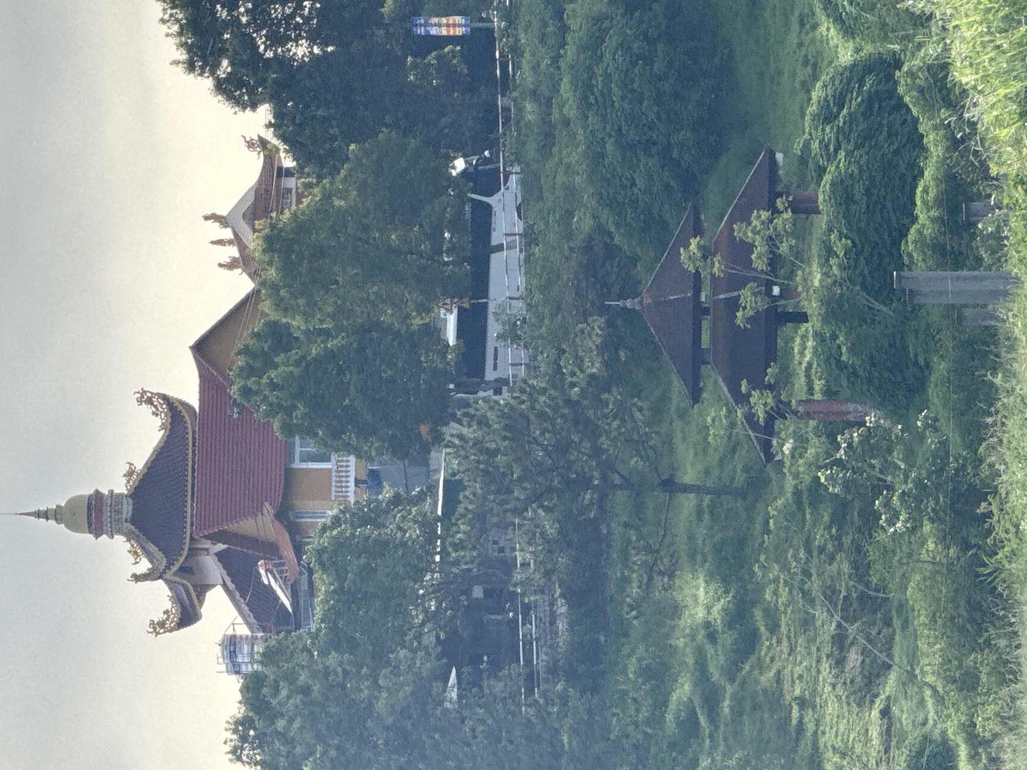 This image captures a serene view of a traditional Vietnamese building, likely a temple or pagoda, situated on a lush green hillside in Da Lat, Vietnam. The building features intricate architecture with a multi-tiered, curved roof adorned with golden dragon motifs, characteristic of Vietnamese design. In the foreground, a small, open-sided pavilion with a similar dark, tiled roof provides a sheltered resting spot amidst the verdant landscape. The surrounding environment is characterized by rolling hills covered in dense vegetation, trees, and grasses, suggesting a natural and tranquil setting. A road runs across the middle ground, where a white bus is visible, indicating the presence of transportation and potential visitors. The sky is overcast, casting a soft, diffused light over the scene. The overall impression is one of peacefulness and natural beauty, with the traditional architecture harmoniously integrated into the landscape of Da Lat.