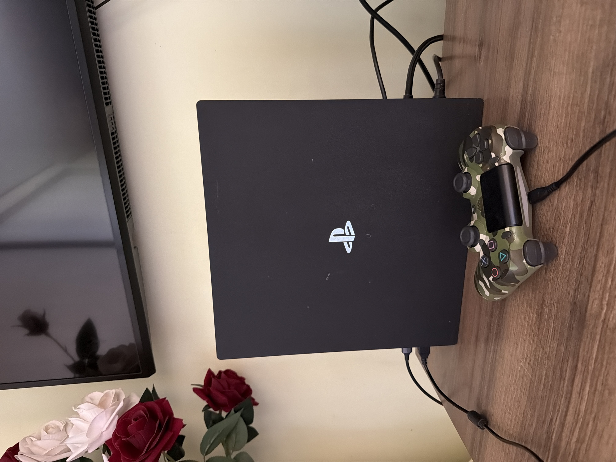 An overhead shot shows a black PlayStation 4 console, with a white PlayStation logo on its top surface, standing vertically on a wooden table. A camouflaged PlayStation controller rests in front of the console. In the background, a television screen reflects a bouquet of artificial roses, red and white, that are positioned to the left of the frame, against a cream-colored wall. The scene appears to be indoors. The city of Samarkand, Uzbekistan is not depicted in the image.