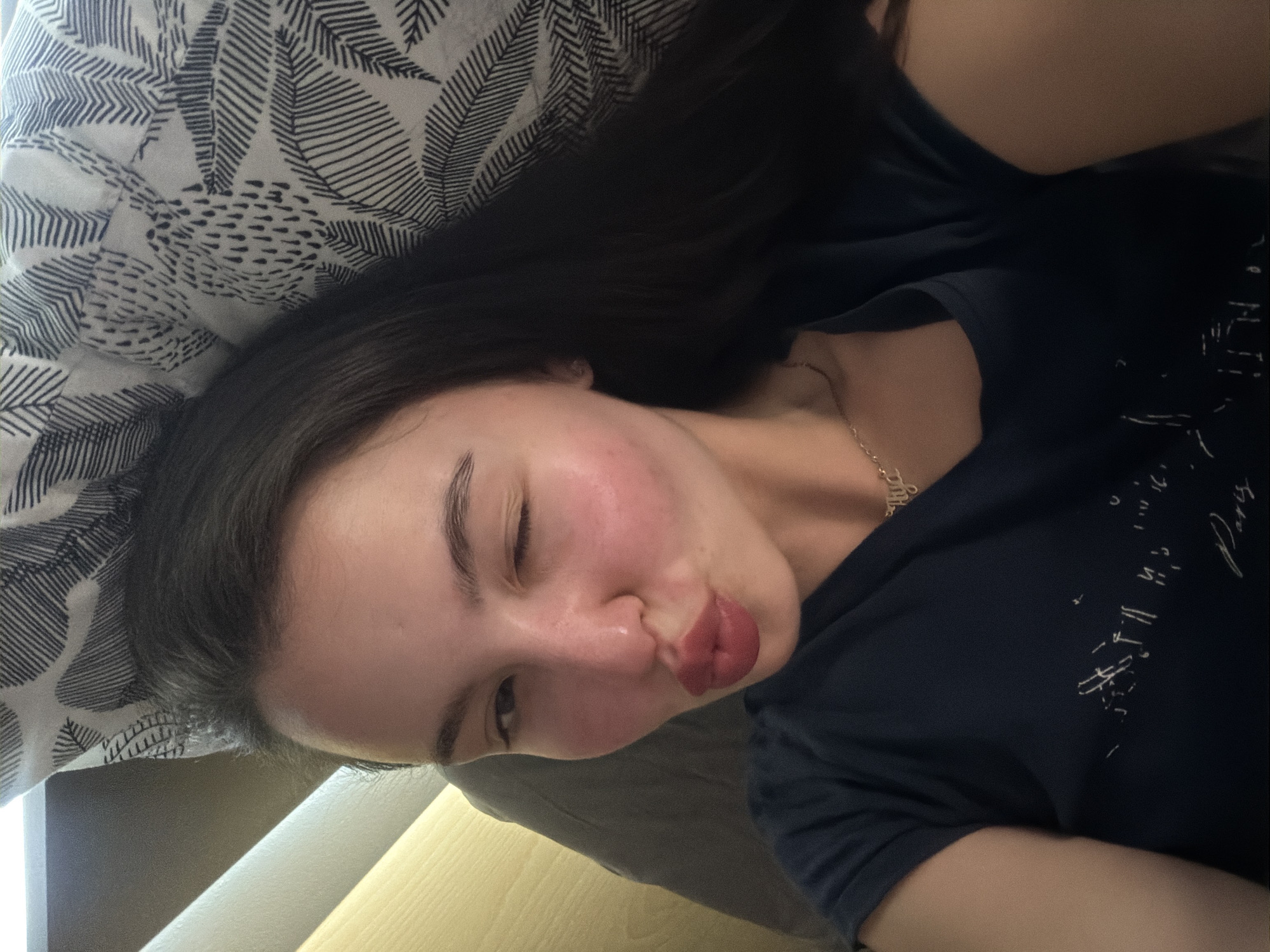 A young woman lies on a bed. Her head rests on a pillow with a black and white leaf-patterned cover. She wears a dark blue t-shirt and a thin gold chain necklace. The room is illuminated by a warm light, and a wooden table is visible in the background. She is pouting her lips as if to kiss the camera. She has one eye closed. The location is in the city of Unknown, Unknown.