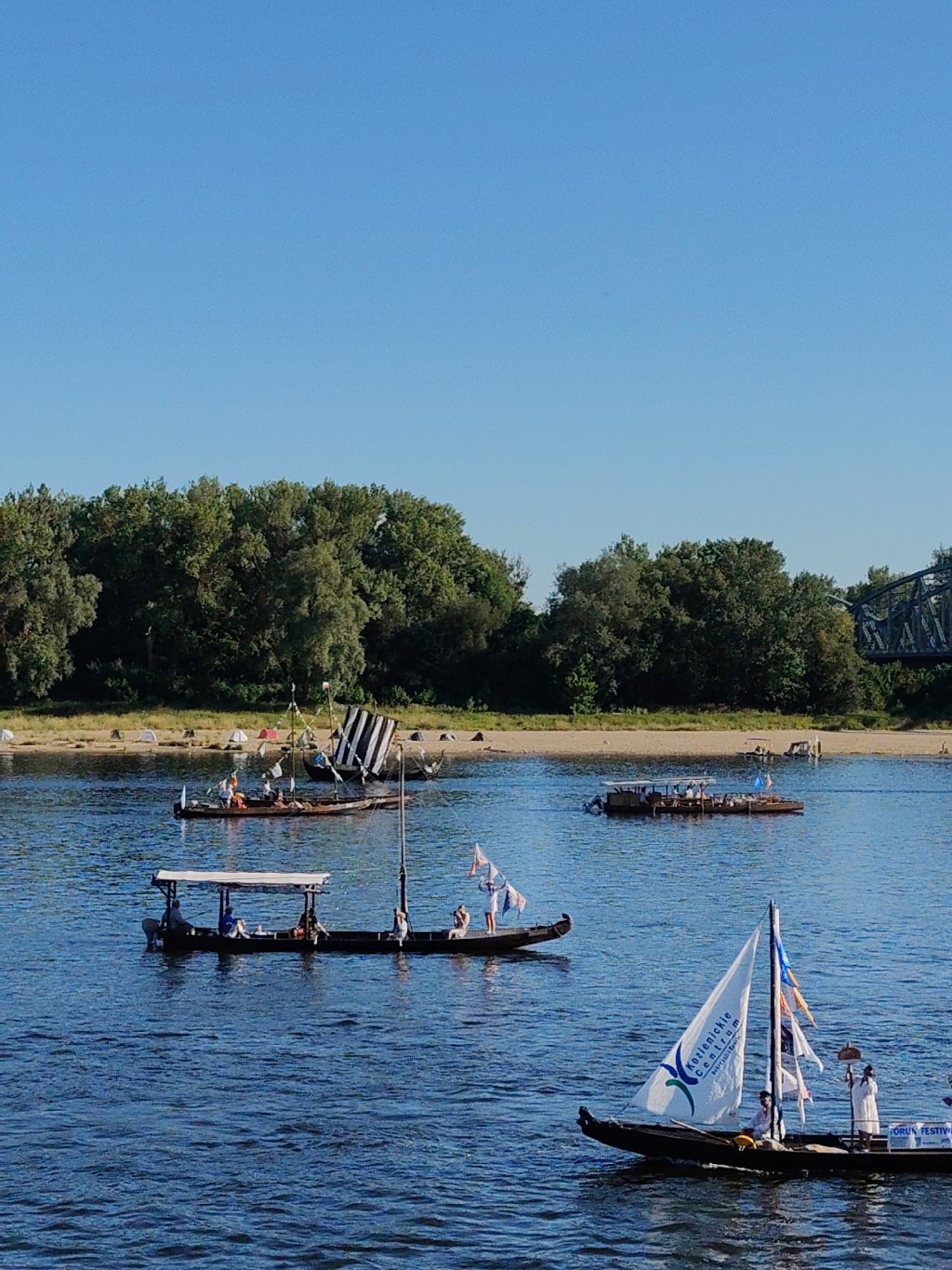 An eye-level, slightly low-angle, medium shot captures a bright, sunny day with a clear blue sky over a wide, blue river. On the river, several boats are navigating. In the foreground, a dark-hulled sailboat with a white sail bearing the logo "Kozienicke Centrum" is sailing from left to right. Two people are on board this sailboat. To the left of the sailboat, a longer, dark-hulled boat with a white canopy is moving from left to right, with several people seated under the canopy. Behind this boat, another dark-hulled boat with a large black and white striped sail is stationary. Further in the distance, a larger boat with a canopy is moving from left to right. The riverbanks are lined with lush green trees. On the near riverbank, in the middle ground, several white tents are set up on a sandy shore. In the far background, a metal bridge is visible, arching over the river.