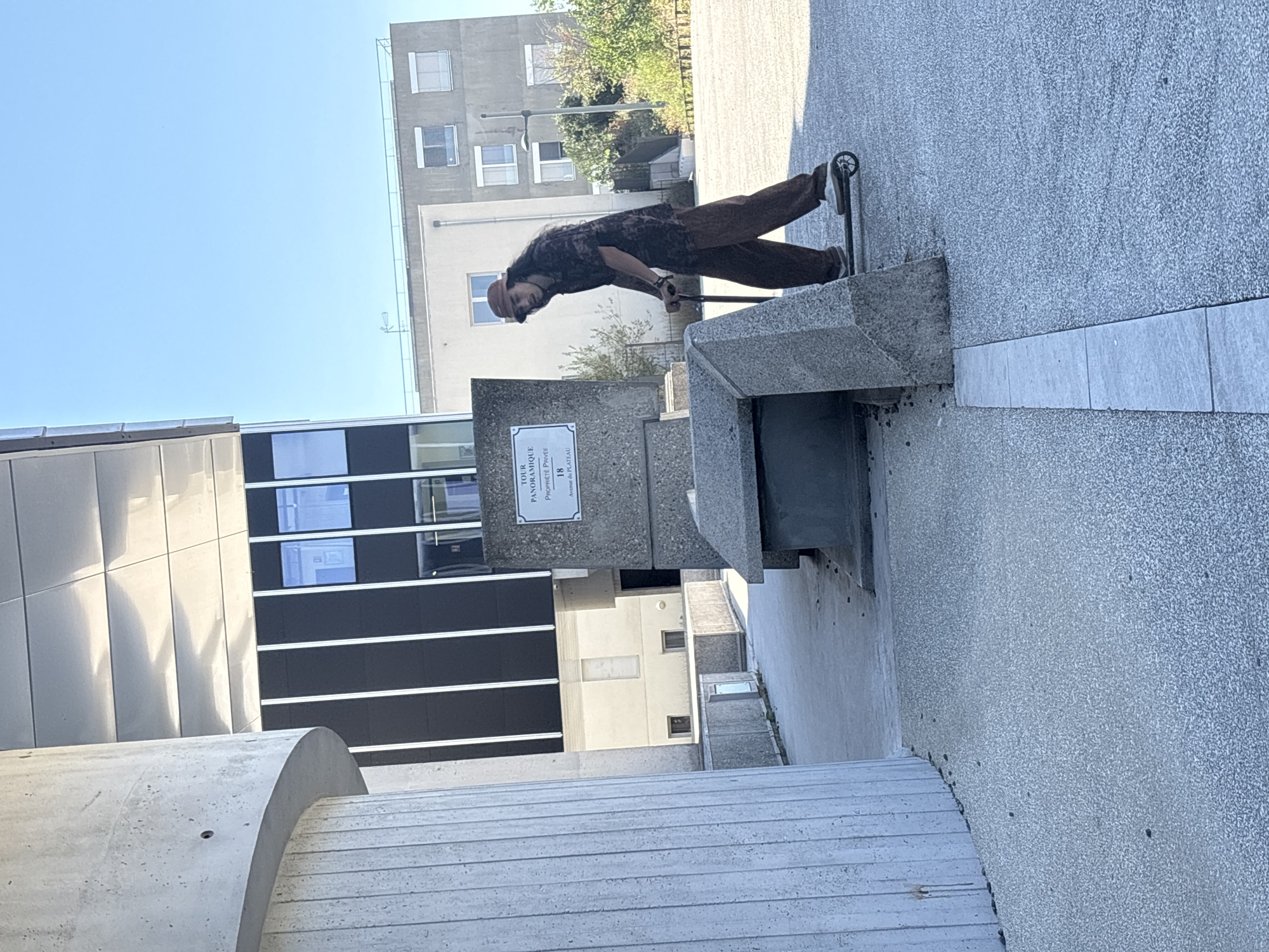 A person rides a scooter past a concrete structure with a sign that reads "TOUR PANORAMIQUE" and "PROPRIETE PRIVEE 18 Avenue du Plateau". The scene appears to be in an urban outdoor setting with modern architecture, including a building with black and white vertical accents and a large concrete pillar. The sky is clear and blue.