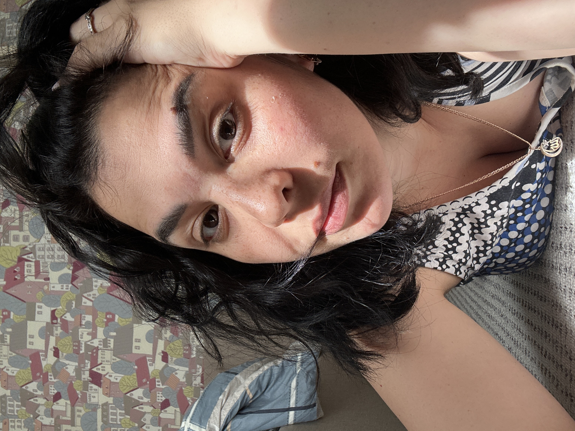 A close-up, slightly angled selfie captures a woman with dark, wavy hair, reclining indoors. She looks directly at the camera with a calm, natural expression and a faint smile. Her right hand, adorned with a ring, rests on her forehead, causing visible wrinkles on her skin. Strong, direct sunlight illuminates her face and shoulder, creating prominent highlights and shadows, suggesting a morning or late afternoon setting.
Her skin shows visible pores, some minor redness, and a small mole near her right eyebrow. A few small droplets of moisture are visible on her forehead and cheek. She wears a patterned top featuring blue and white polka dots mixed with black and white stripes. Around her neck is a gold-toned necklace with a circular charm, which appears to have a stylized symbol or design, possibly an animal or emblem, surrounded by small decorative elements.
The background behind her head features a patterned surface, possibly wallpaper or fabric, with colorful, stylized depictions of small buildings and trees. A grey knitted blanket or throw and a striped blue and white pillow are visible, suggesting a cozy, domestic indoor setting such as a bed or sofa. No discernible text is visible in the image.