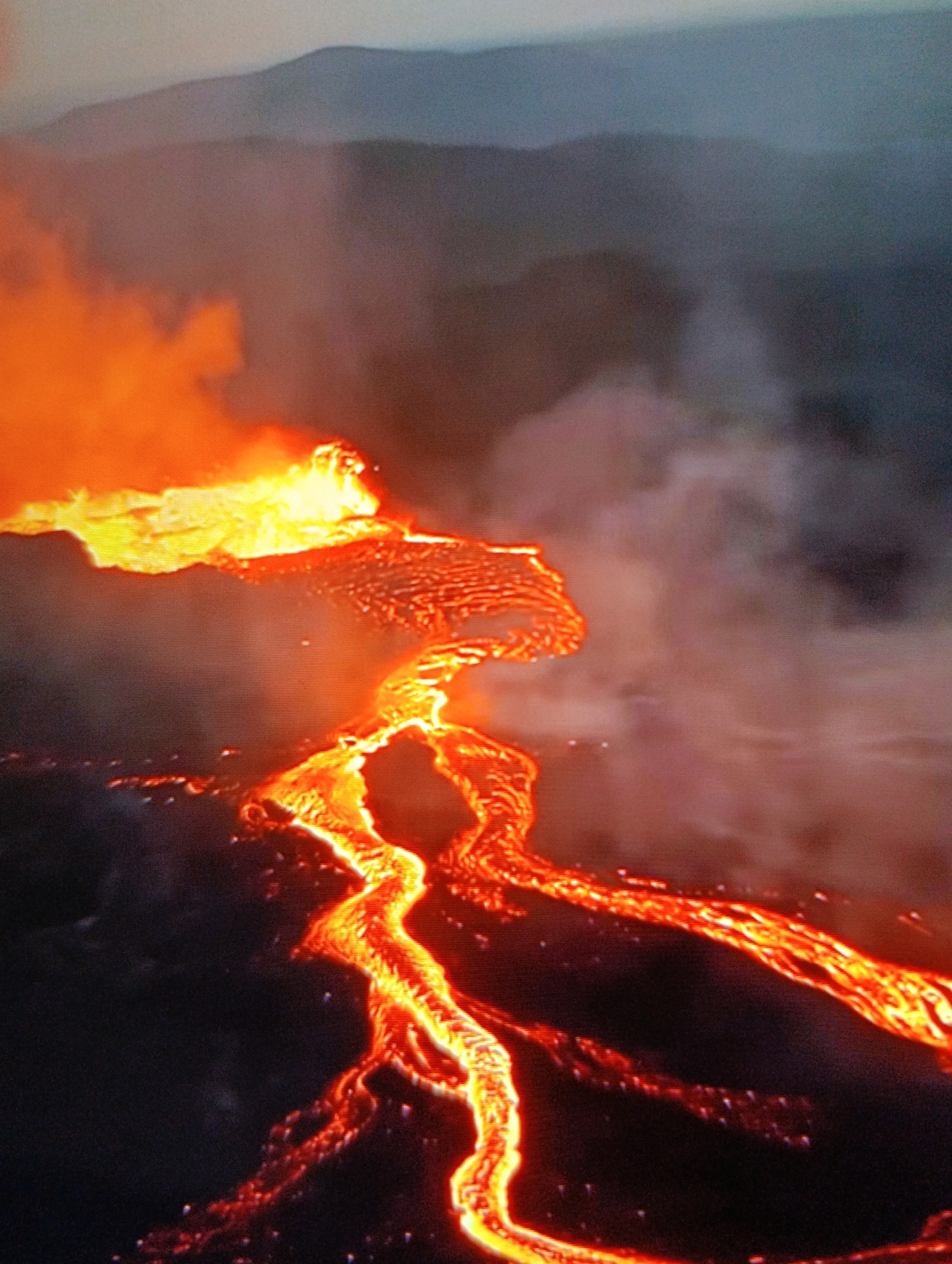 The image depicts an active volcanic eruption. Molten lava flows downhill, glowing orange and yellow, against a dark background. The lava originates from a fiery area at the top left of the image. Smoke and ash are present in the air. Hills are visible in the background. The city location is Unknown, Unknown.