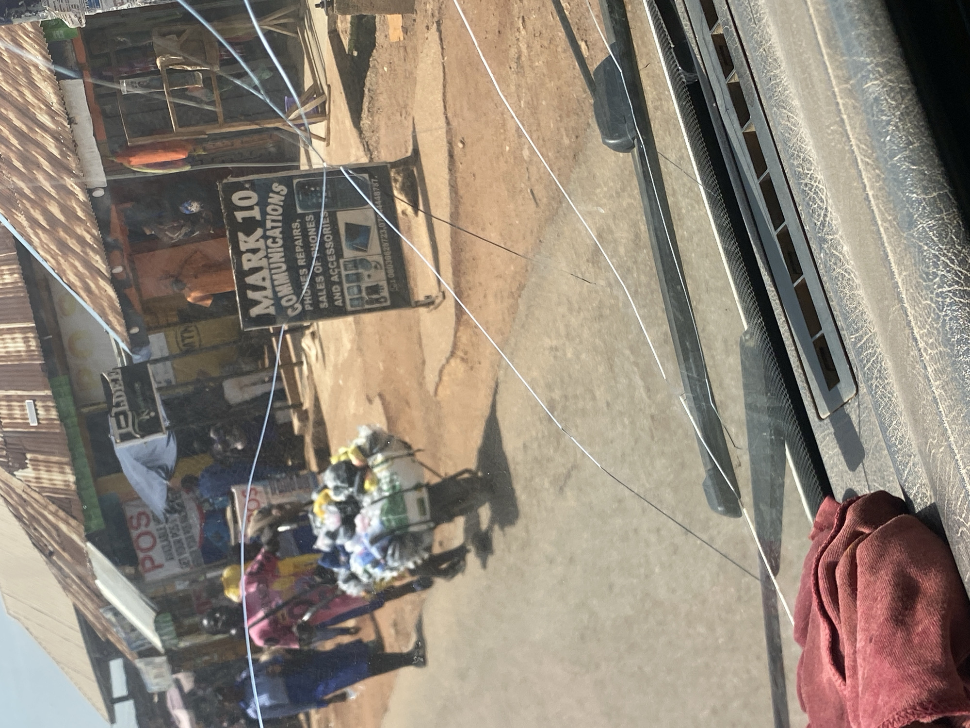 The image depicts a bustling street scene in Jos, Nigeria. In the foreground, a red cloth lies on the dashboard of a vehicle, with what appears to be a cracked windshield obscuring the view. Beyond the windshield, a street vendor pushes a cart laden with assorted goods, including clothing and other items. Pedestrians are visible walking along the street, and shops line the side of the road, with signs indicating services such as "POS" and "MARK 10 COMMUNICATIONS" offering phone repairs and sales. The environment suggests an active marketplace with a mix of commercial activity and daily life.