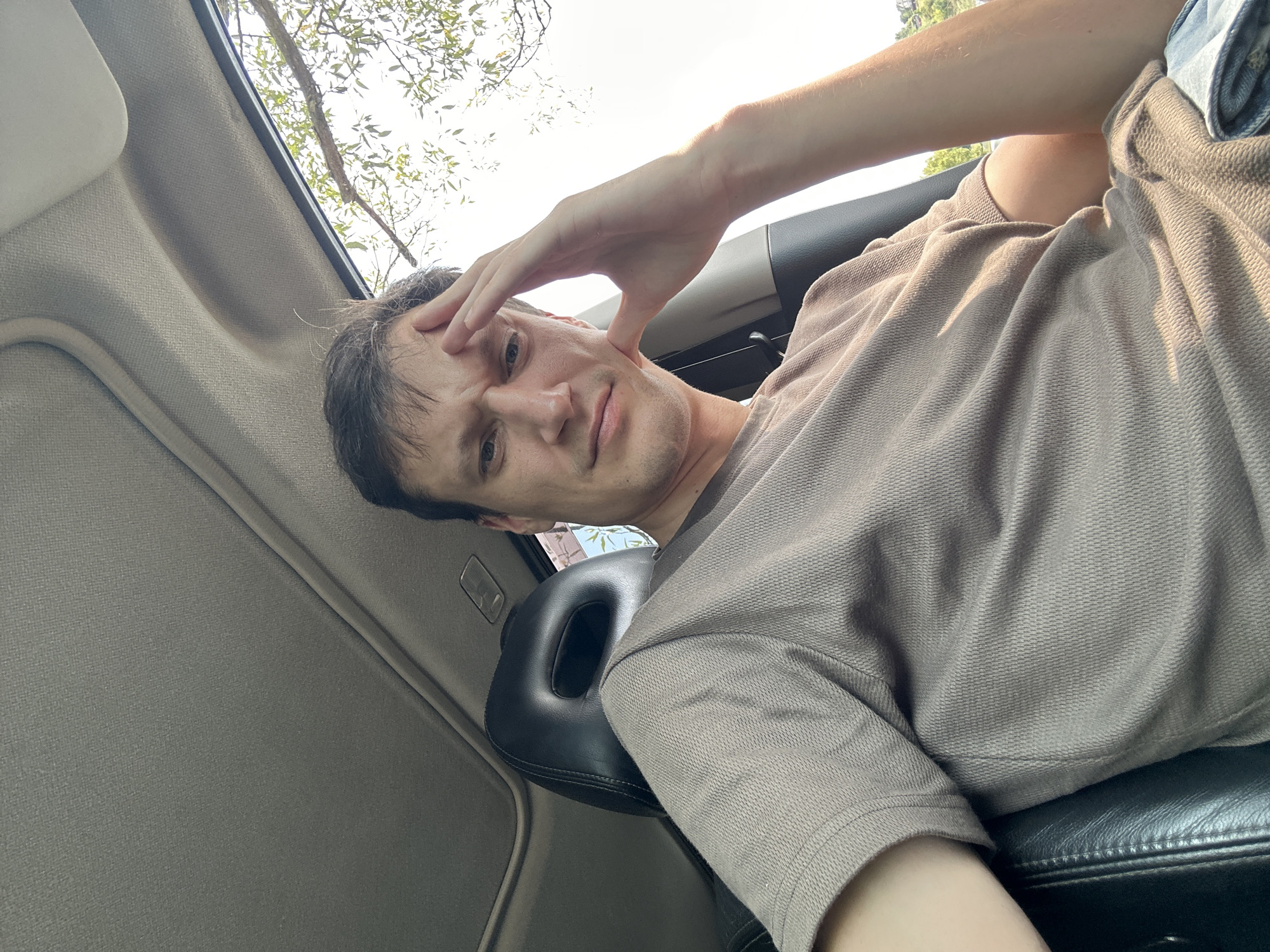 A man sits in the driver's seat of a car, facing the camera. He has short, dark hair and is resting his right hand on his forehead, his gaze directed at the lens. He is wearing a taupe-colored t-shirt. The interior of the car is visible, including the headrest of the passenger seat and a sunroof. Outside the car, sunlight filters through tree branches. The scene appears to be captured in an outdoor setting, possibly a road or parking area.