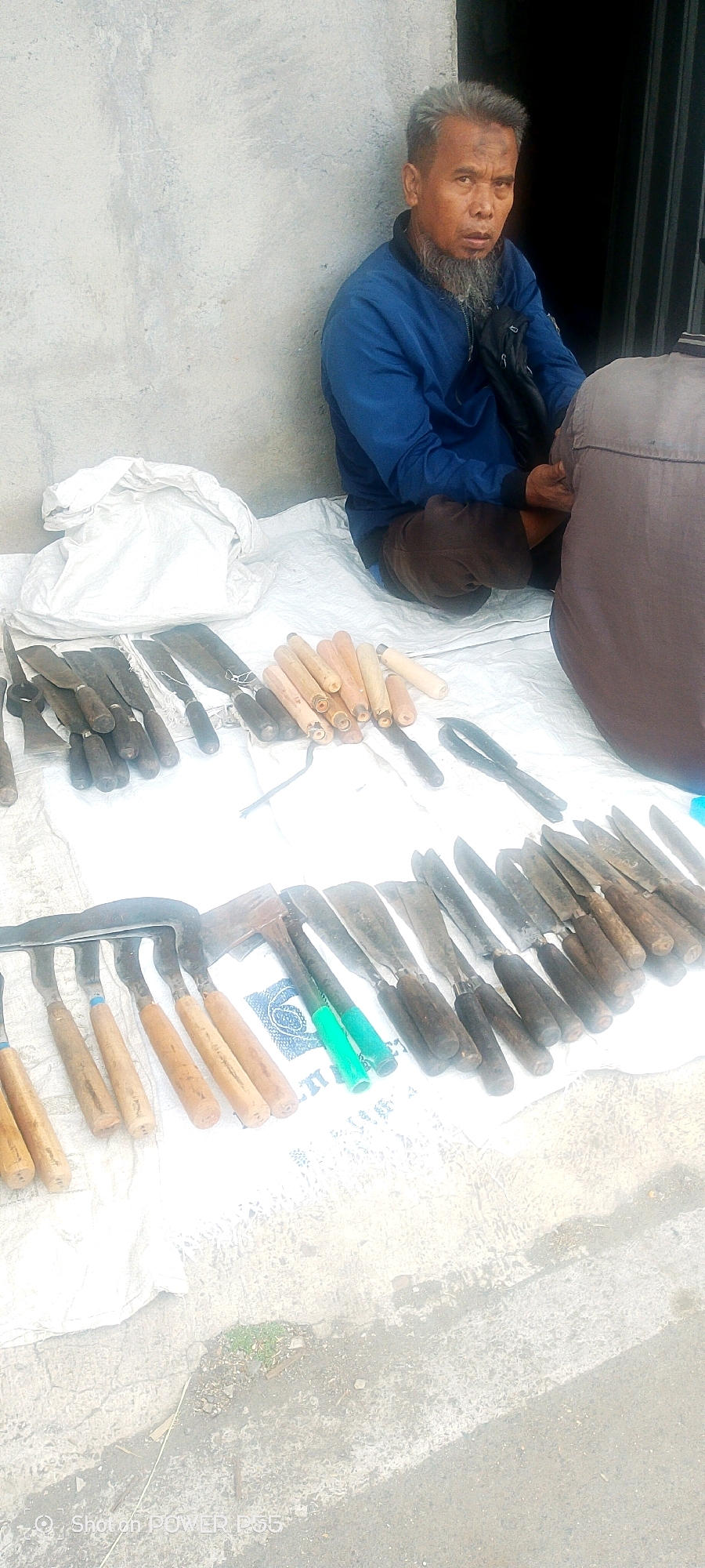 An elderly man with graying hair and a beard, wearing a blue jacket and brown pants, sits on a white cloth laid on the ground. He is looking directly at the camera. To his left, arranged on the white cloth, are numerous tools with wooden handles, including knives and what appear to be agricultural implements. A white sack is placed behind the man. Another person, whose face is not visible, is partially seated to the right of the man, wearing a brown garment. The background consists of a concrete wall and a dark opening, possibly a doorway. The scene appears to be outdoors on a paved surface.