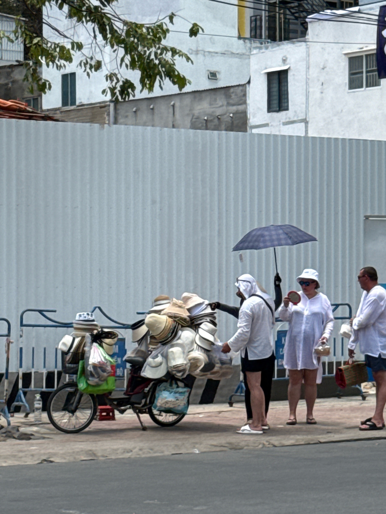 The image depicts a street scene in Nha Trang, Vietnam, possibly near the coast. A motorcycle is laden with numerous hats, suggesting a vendor's stall. Three individuals are visible interacting near the motorcycle. One person, dressed in a white thobe and headscarf, appears to be engaged in a transaction or conversation. Another individual, wearing a white hat and dress, holds a small round object and an umbrella. The third person, a man in a white shirt and sunglasses, stands to the right. The environment includes a grey corrugated metal fence, buildings in the background, and a street in the foreground. The overall impression is one of a busy marketplace or street commerce.