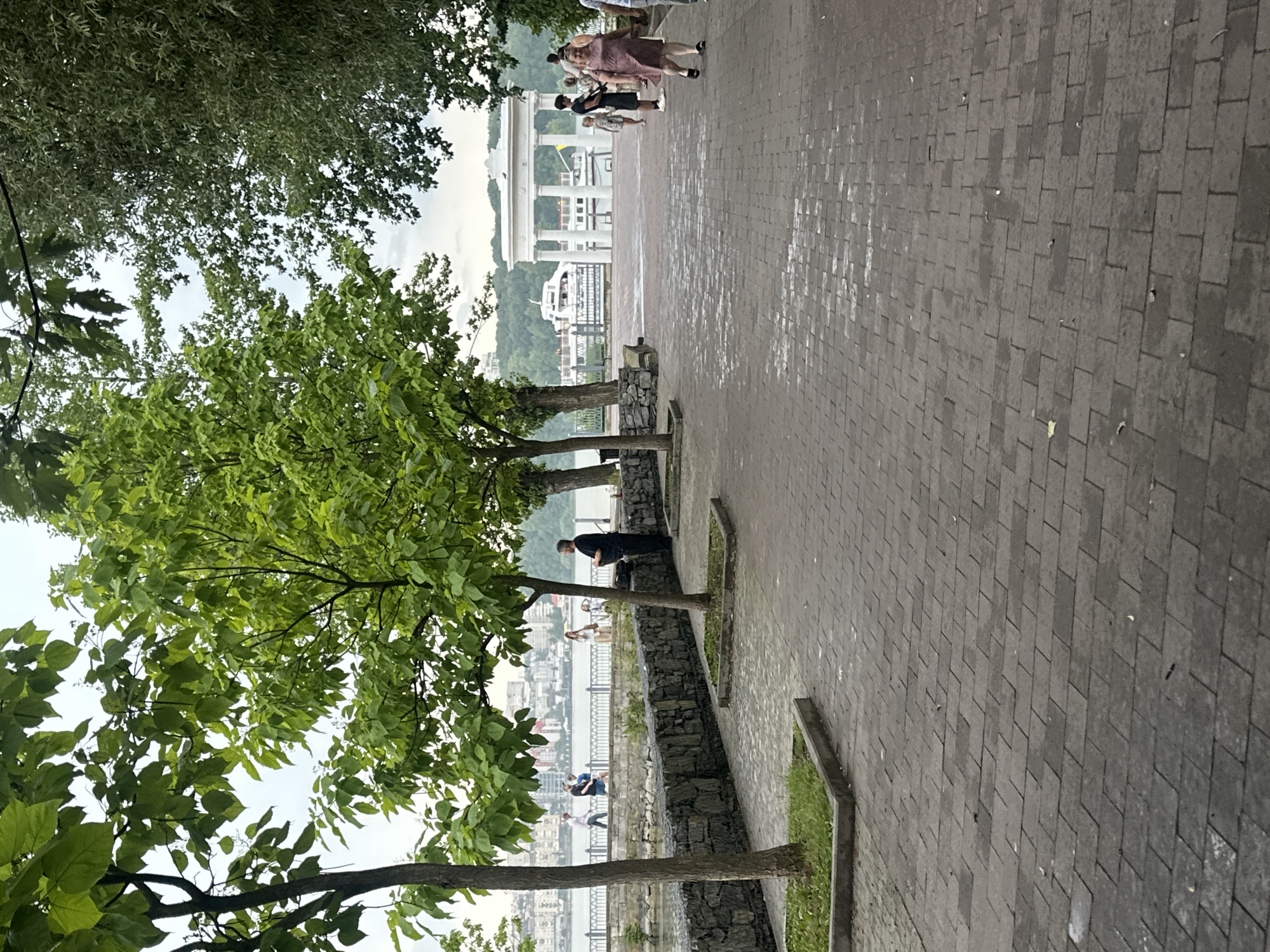 The image depicts a waterfront promenade in Ternopil, Ukraine, likely near the Ternopil Pond. The foreground is dominated by a paved walkway made of dark grey bricks, showing some signs of wear and moisture. Several young trees with vibrant green leaves are planted in square, concrete-lined beds along the walkway's edge. To the left, a stone retaining wall separates the walkway from the water, which is visible in the background. Across the water, the cityscape of Ternopil is discernible, with buildings and a distant hillside covered in trees.
Further along the promenade, a white, classical-style gazebo or rotunda stands near the water's edge, with a pleasure boat docked beside it. Several people are seen walking and standing on the promenade. In the mid-ground, a man in dark clothing stands near the stone wall, facing away from the camera. To the right, a group of people, including a woman in a pink dress, are walking away from the camera. The sky is overcast, suggesting a cloudy day. The overall impression is that of a public park or recreational area by a body of water.
