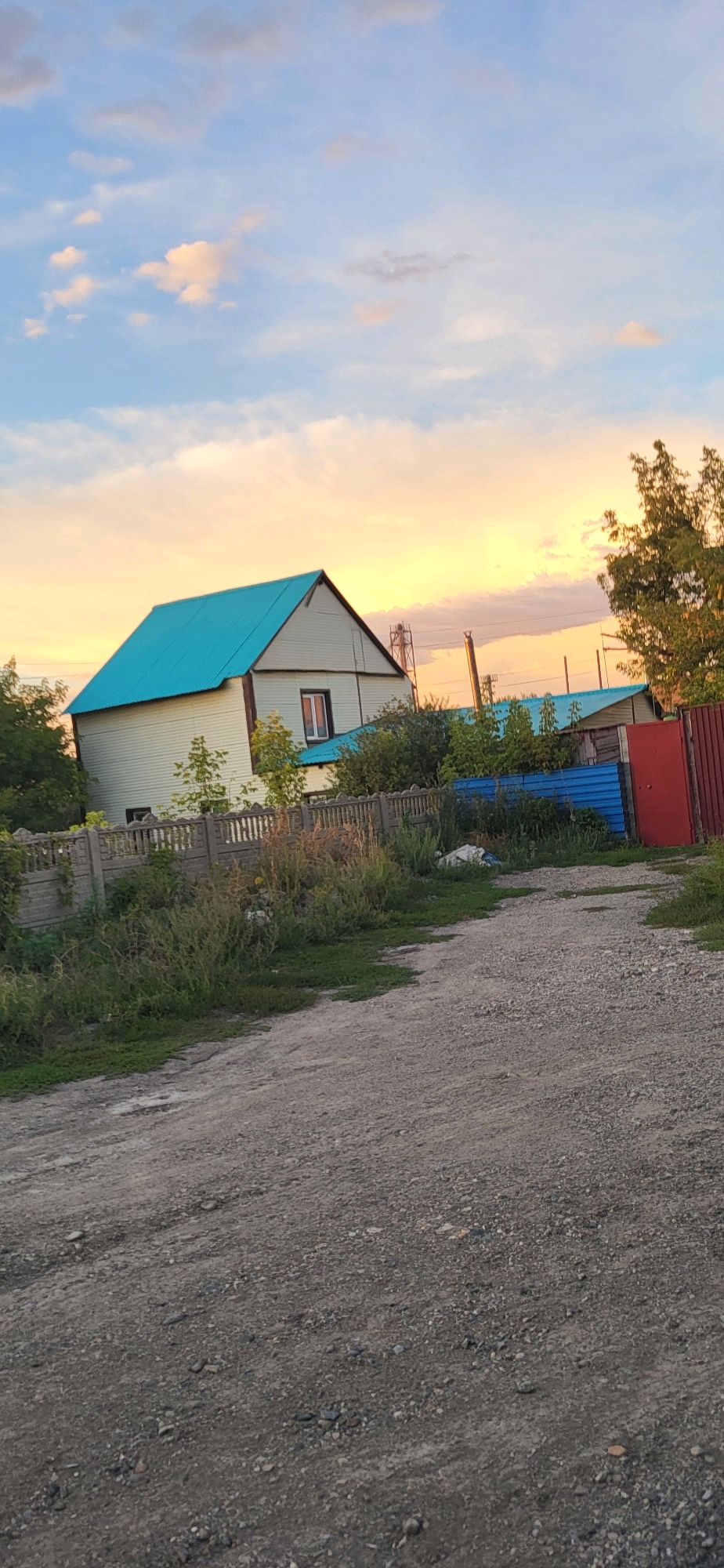 A dirt road leads through a rural area with a two-story house that has a bright turquoise roof. The house is light beige with dark trim around the windows. There is a window on the second floor with an orange glow coming from inside. The house is bordered by a concrete fence and overgrown vegetation. Beyond the fence, there are several other structures with turquoise roofs and a red metal fence. In the background, trees and power lines are visible, and the sky is a pale blue with scattered clouds tinged with orange and pink from the sunset.