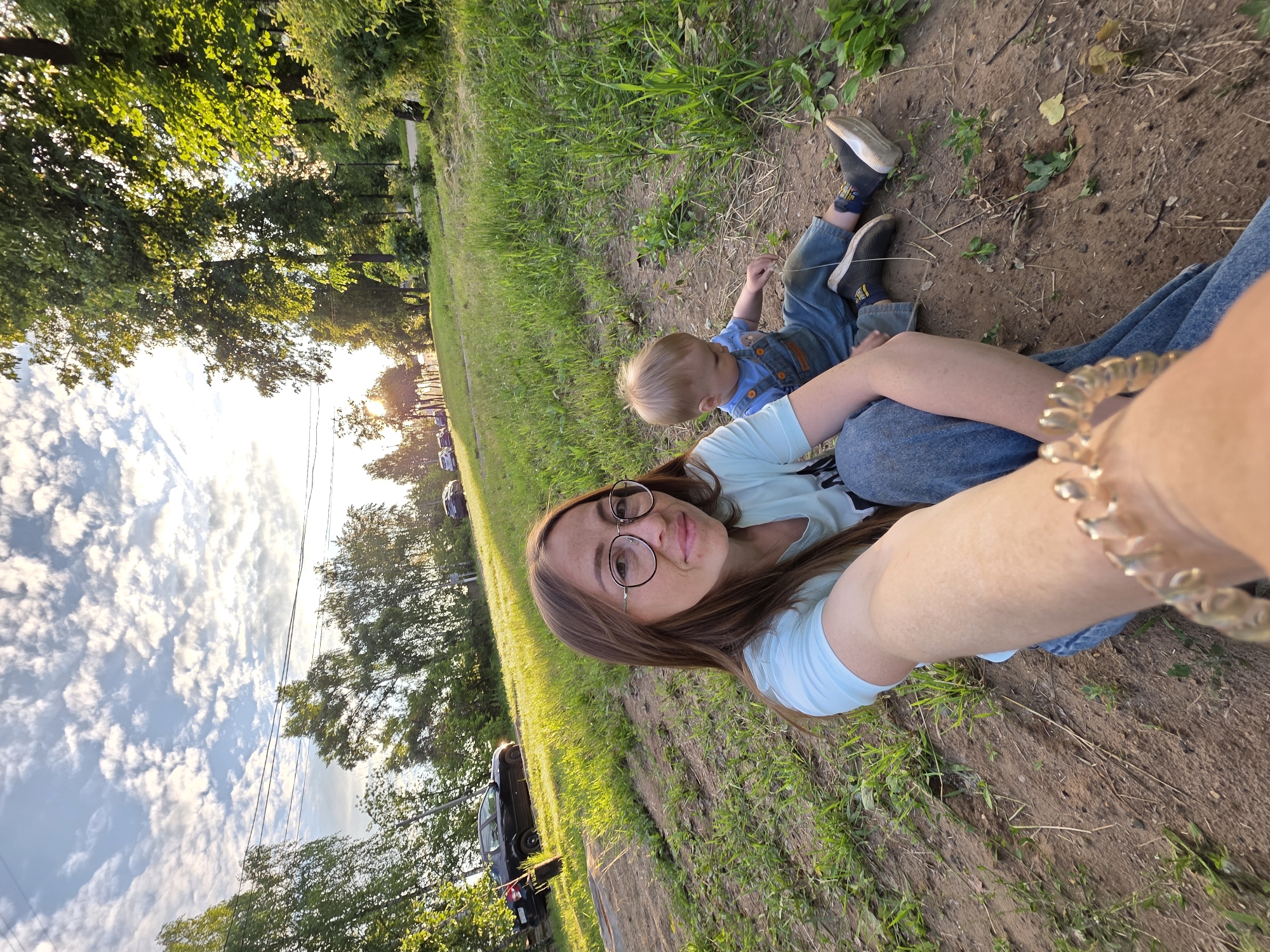 This full-frame, eye-level selfie captures an adult female and a young child situated on a dirt and grass incline. The image is taken outdoors during daylight hours, characterized by a bright, partly cloudy sky and prominent sun glare from the upper right, indicating either sunrise or sunset.
The adult female, positioned in the foreground and center, has long, straight brown hair, wears round-framed glasses, a light blue t-shirt, and blue jeans. Freckles are visible on her nose and cheeks. She is looking directly at the camera, holding it with her left hand, which displays a gold-toned bracelet with large, interlocking links in the lower right foreground. She is sitting on the ground with her legs bent.
To the adult's right and slightly behind, a young child with light blonde hair is seated on the ground, wearing a blue t-shirt with a bear graphic and blue denim overalls. The child is looking down and away from the camera, seemingly focused on the ground or a small plant in front of them. Dark sneakers with white soles are visible on the child's feet.
The ground consists of dry dirt interspersed with patches of green grass and sparse weeds. The terrain slopes upward towards the background. In the mid-ground, a paved road with several parked cars (including a black sedan on the left) is visible. Beyond the road, numerous deciduous trees with green foliage line the horizon, suggesting a park or green belt area. Overhead, power lines stretch across the sky in the upper left quadrant.
While the image depicts an outdoor recreational area, no specific architectural features, landmarks, or signs are discernible to definitively pinpoint the exact location within the city of Chaikovsky, Russia.