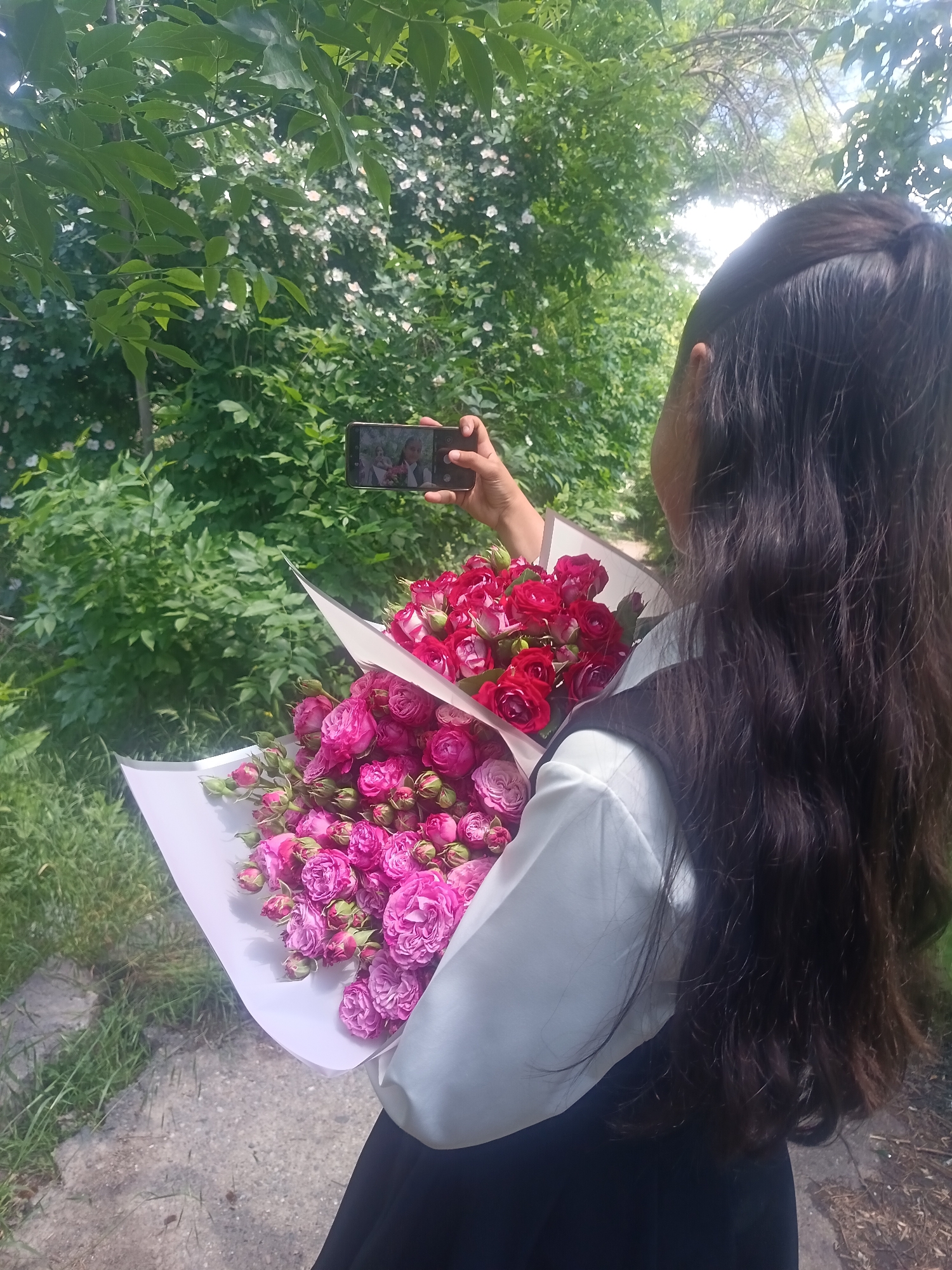 A young woman with long, dark hair, wearing a white shirt and black skirt, stands outdoors and takes a photo of two bouquets of roses with her smartphone. One bouquet is composed of vibrant pink roses and the other of red roses. The background consists of lush green foliage, with a light-colored path or walkway visible in the foreground.