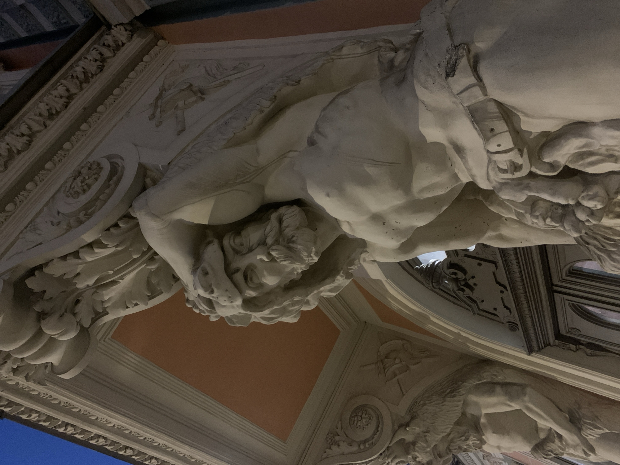 The image shows a close-up of the exterior architecture of the Singer Building in Saint Petersburg, Russia, at twilight. The building features ornate white stone sculptures, specifically two muscular figures that appear to be telamones supporting an archway. The figure in the foreground is depicted with a lion's pelt draped over its head and shoulders, and a belt around its waist. The sculpture exhibits detailed musculature and facial features, including a beard. In the background, a similar sculpture of a figure with a fur pelt is partially visible. Above the sculptures, the building's facade has decorative carvings and a salmon-colored ceiling. The sky is a deep blue, indicating it is either early morning or evening. The overall scene conveys a sense of grandeur and historical architecture.