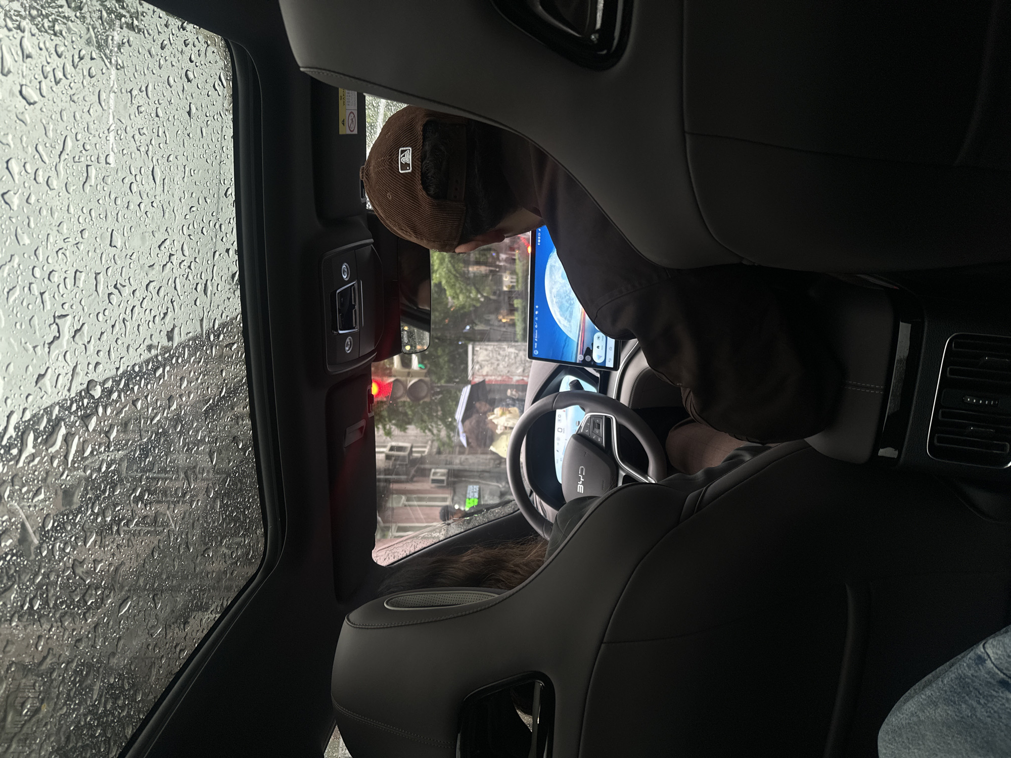 The image shows the interior of a car, likely a BYD brand given the steering wheel logo, during a rainy day. Raindrops cover the window. A person wearing a brown baseball cap is in the driver's seat. The car's infotainment screen is visible. The scene appears to be in Yerevan, Armenia, identifiable from the architecture and streetscape visible through the front windshield. The car is stopped at a traffic light.