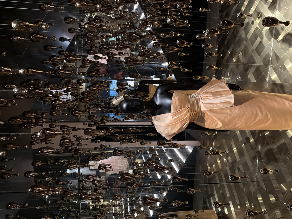 This image captures an interior scene at the Galerie Dior in Paris, France, within an exhibition space characterized by opulent display and reflective surfaces.
In the foreground, slightly right of center, stands a black, headless female mannequin. It is dressed in an elegant, one-shoulder, floor-length gown of a light peach or champagne color, crafted from a lustrous, draped fabric, featuring a ruched bodice and a prominent peplum detail at the waist.
The environment is dominated by an art installation consisting of thousands of gold-colored, teardrop-shaped objects suspended from the ceiling at varying heights, creating a dense, shimmering canopy. The walls and floor are clad in highly polished, mirrored surfaces, which generate multiple, complex reflections of the hanging objects, the mannequins, and the architectural features of the room.
Through these reflections, a male individual is visible on the right side of the frame. He has a beard, wears a dark shirt, and is holding a smartphone, indicating he is actively photographing the scene. Further reflections reveal another mannequin in the background, displaying a light-colored, intricately detailed gown (possibly pink), and a modern staircase with illuminated risers. The lighting is ambient, accentuating the reflective properties of the installation and the fabric of the displayed garments.