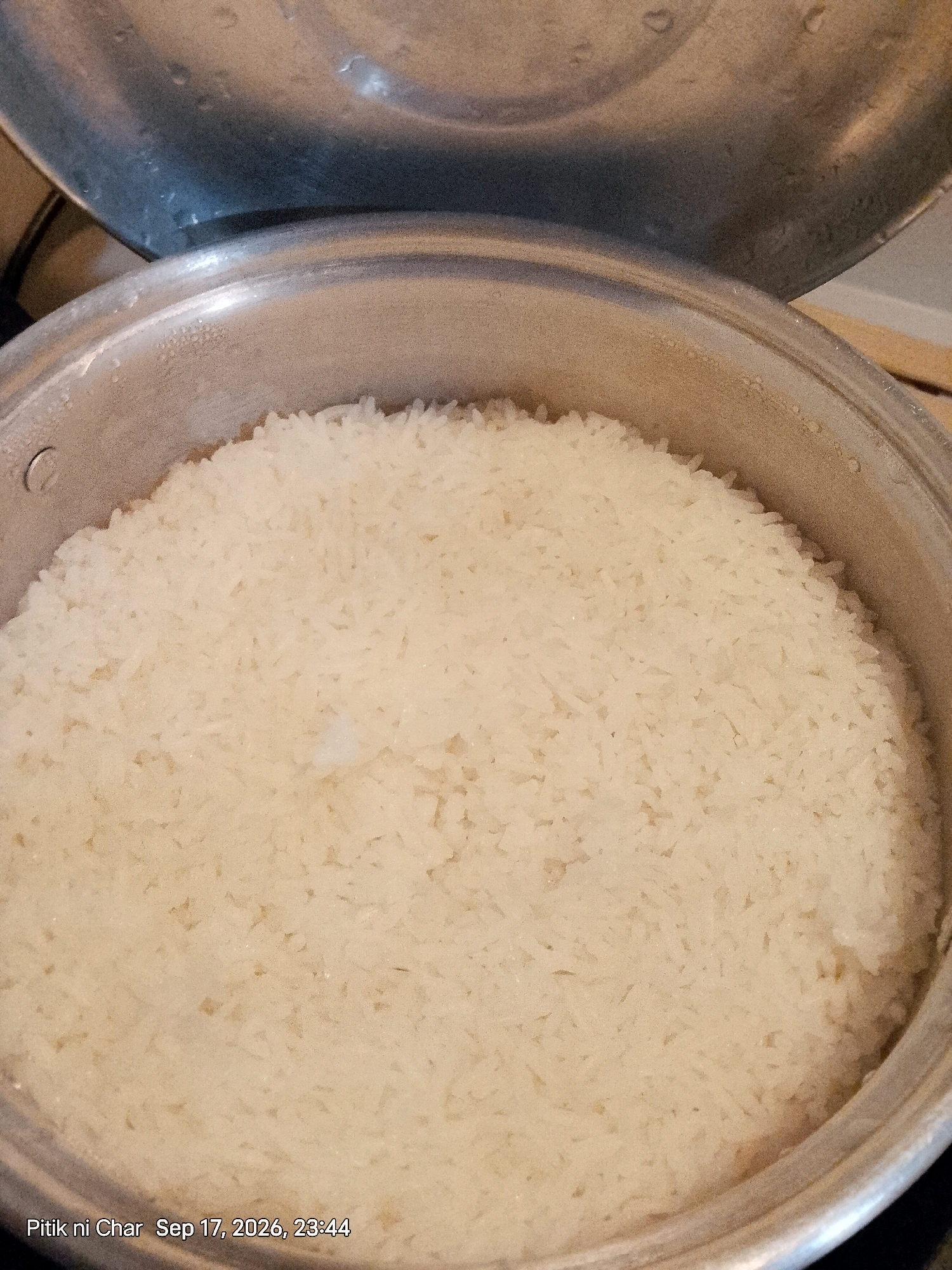 A top-down, close-up shot of freshly cooked white rice fills the frame of a pot. The rice appears fluffy and is piled high, with some grains showing slight browning at the edges, suggesting it may have been steamed or lightly fried. The pot itself is made of silver-colored metal, with a slightly worn appearance and visible condensation on its rim. An open lid, also silver-colored, is positioned above and behind the pot, allowing a view into the cooked rice. The background is blurred, but hints of a wooden surface and what appears to be a dark countertop are discernible. The bottom left corner of the image contains a timestamp and a caption: "Pitil ni Char Sep 17, 2026, 23:44". This image is not in Davao City, Philippines.