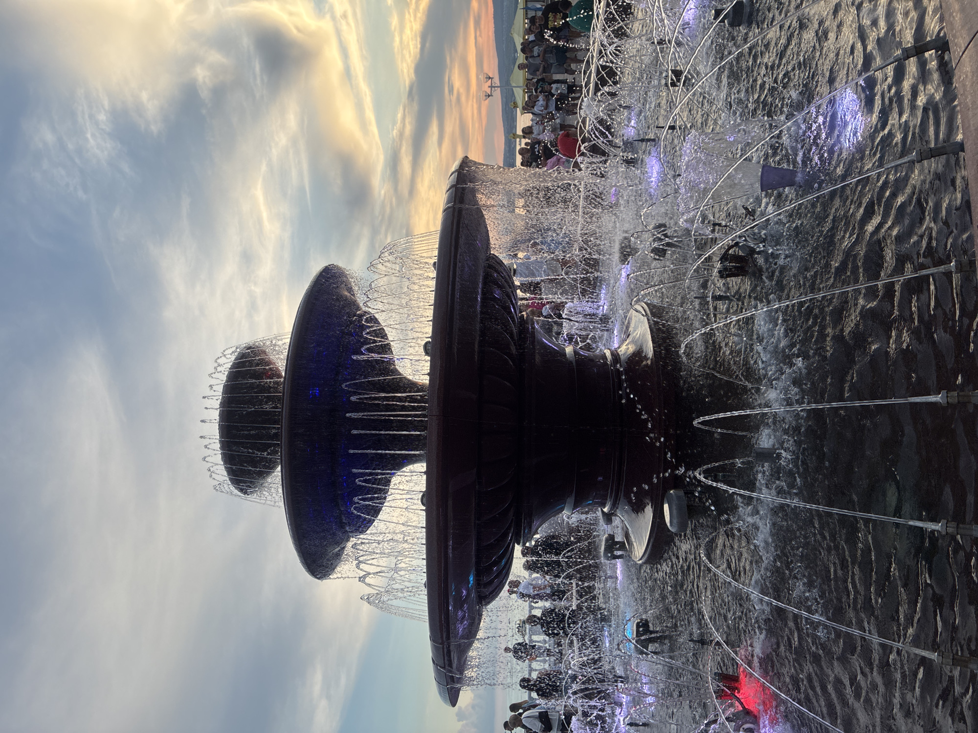 A large, tiered fountain is the central focus of the image, with water cascading down its sides and spraying upwards from a surrounding pool. The fountain appears to be made of dark, polished material. Several people are visible in the background, standing near the fountain and along a walkway. They are casually dressed and appear to be enjoying the evening. The sky above is filled with dramatic clouds, illuminated by the warm hues of a sunset, suggesting a coastal or waterfront location. The overall impression is of a public space, possibly a park or promenade, at dusk.