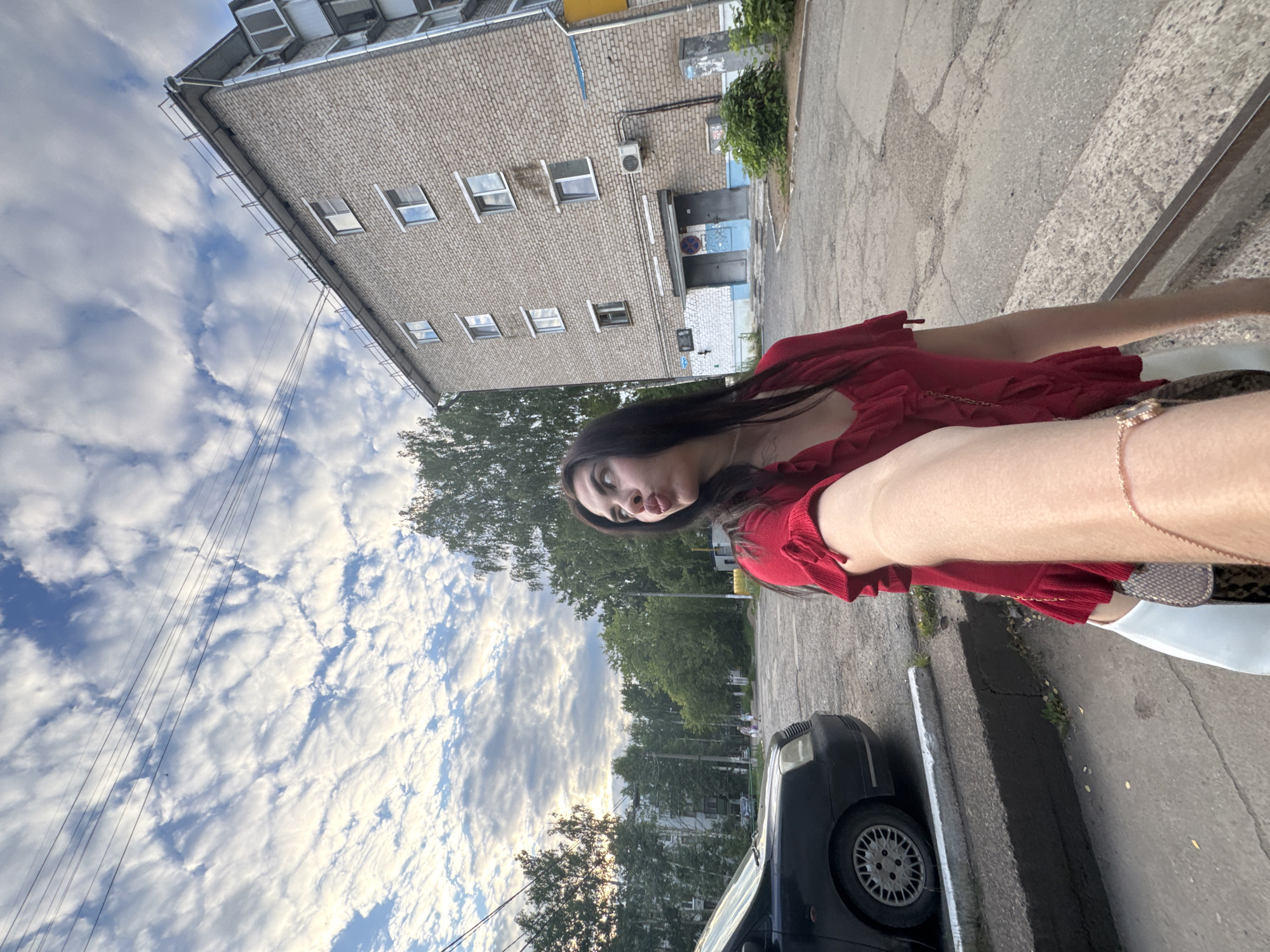 A young woman with long dark hair is posing for a selfie outdoors. She is wearing a red ruffled top and a white skirt. She is looking up and to the right, with her lips puckered. In the background, there is a brick apartment building, a black car, and trees. The sky is cloudy with a blue sky peeking through. The scene appears to be in a residential area in Chaikovsky, Russia.