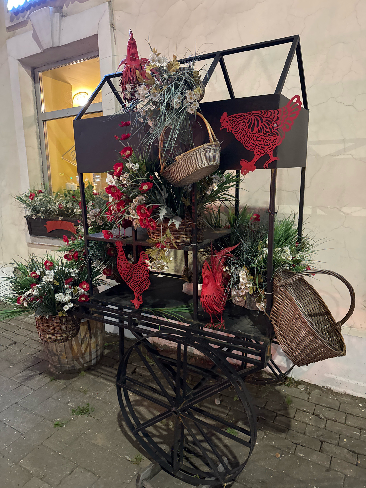 This is a photograph of a decorative cart with floral arrangements and rooster figures, located outside a building on a cobblestone street. The cart is made of black metal and has multiple shelves, a large spoked wheel, and a basket attached to the side. Red roosters, crafted from a thin material, are attached to the cart's structure. Floral arrangements, consisting of red and white flowers and green foliage, are placed in baskets and planters on the shelves and at the base of the cart. A wooden barrel with painted designs is partially visible beneath one of the planters. The building in the background has a window with a yellow light illuminating the interior. The scene appears to be set at dusk or night, as indicated by the lighting. The overall impression is one of a festive or seasonal decoration. The location is stated to be in the city of Tver, Russia. There are no people or animals present in the image.