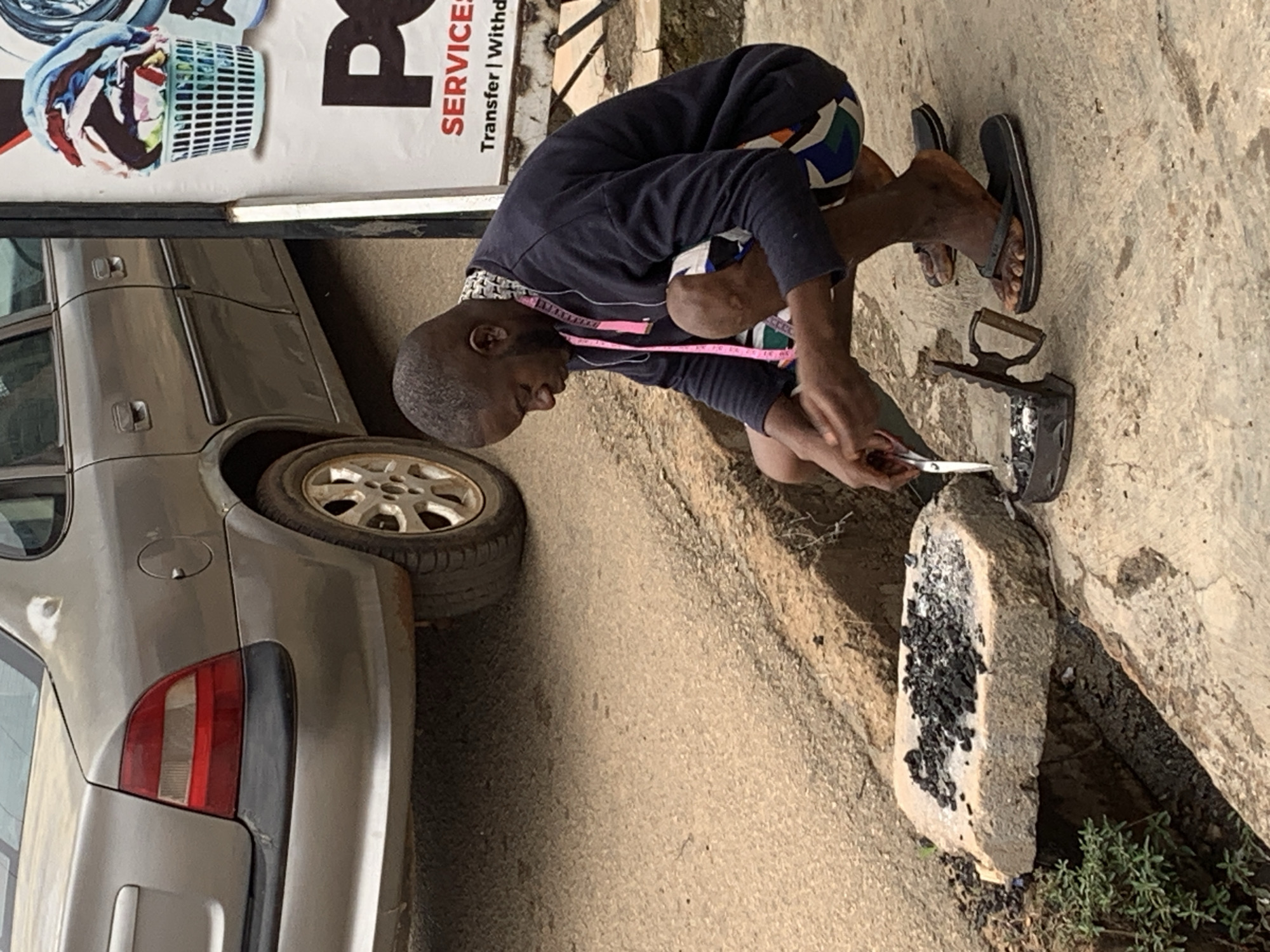 In Jos, Nigeria, a young Black male is seen squatting on a concrete surface next to a roadside. He is wearing a dark blue long-sleeved sweatshirt and colorful shorts. Around his neck, he has a pink measuring tape. His focus is on a charcoal-heated iron, which rests on a flat stone. The iron is positioned over a small pile of glowing embers and charcoal. He is holding a pair of scissors in his right hand, appearing to be tending to the heat source or preparing to use the iron. The background features the rear of a silver sedan and a portion of a poster advertising laundry services, with a basket overflowing with colorful clothes prominently displayed. The ground is a mixture of asphalt and concrete, with a shallow trench running alongside the road.