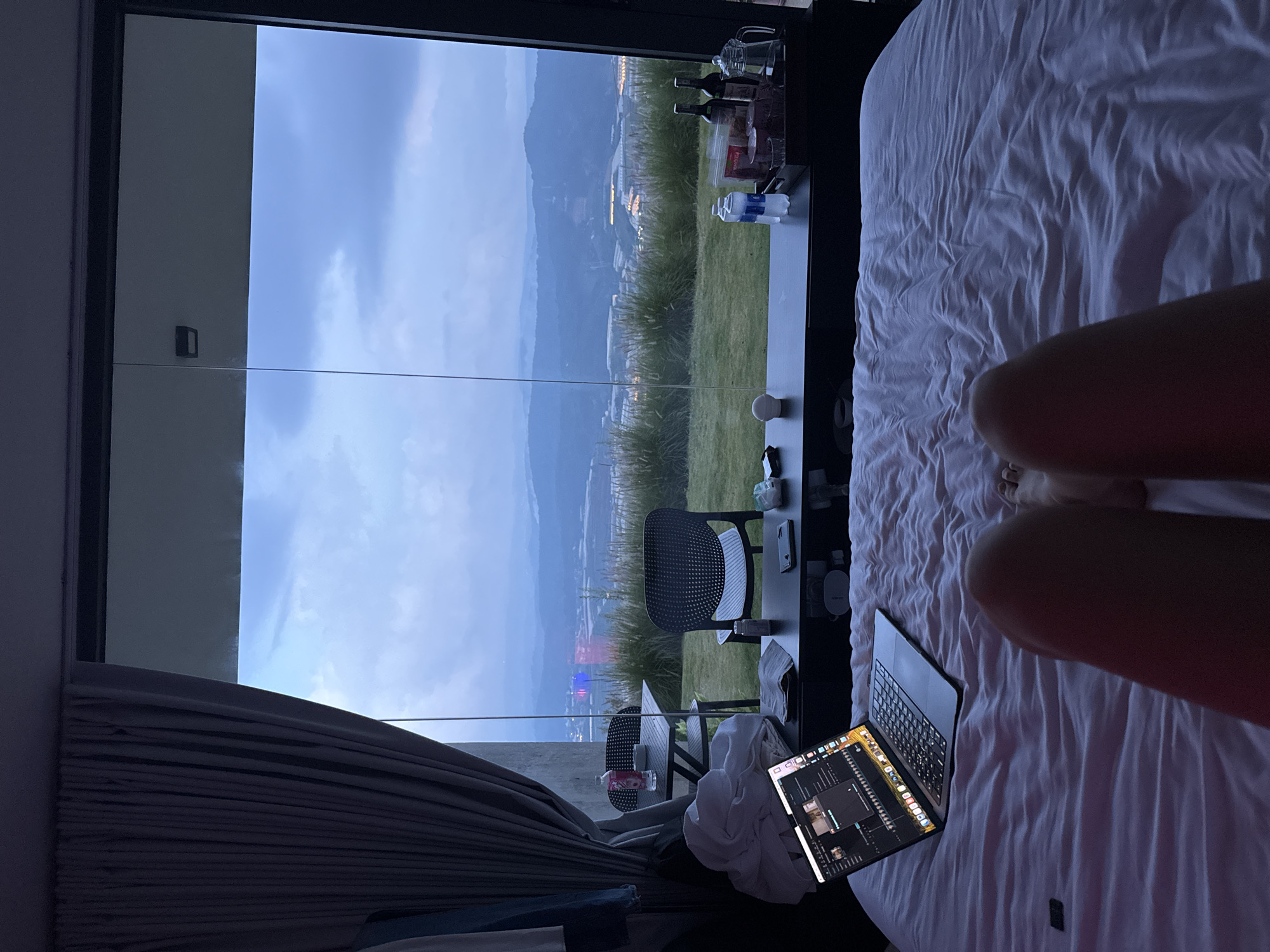 A first-person perspective shot shows a person lying in a bed facing a large window with a view of a city and hills at dusk. The room has a large window that takes up most of the wall, offering a panoramic view of the landscape outside. A laptop is open on the bed in front of the person, displaying a video editing software interface. To the left of the bed, a dark grey curtain is drawn partially across the window. On a low black table in front of the window, there are various items including bottles of water, snacks in packets, two bottles of wine, and a glass pitcher. Further outside the window, on a grassy area, are two chairs and a small table with a water bottle and an ashtray. The city lights are visible in the valley below, and the sky is overcast. The scene appears to be from a hotel room or a rental property in Đa Lộc, Vietnam, given the visual cues of the landscape and architecture. The person's legs and feet are visible at the bottom of the frame.