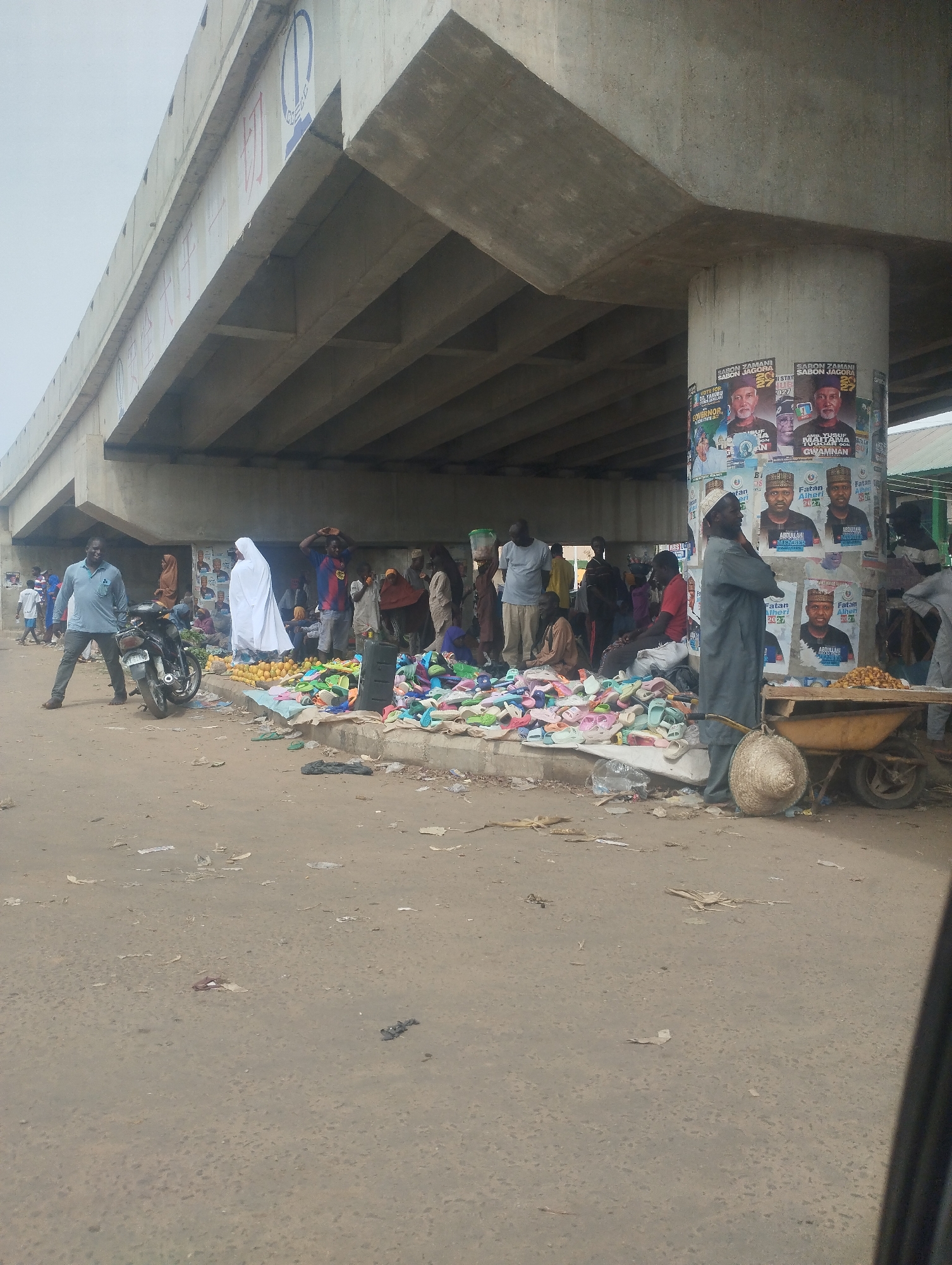 The image shows a bustling informal market taking place under a concrete overpass in Bauchi, Nigeria. A large group of people are congregated around several makeshift stalls and spreads of goods. The primary merchandise visible includes a variety of colorful plastic sandals and a selection of citrus fruits. A man in a grey robe stands near a wheelbarrow, a woven hat resting on its side. Another man in a light blue shirt is standing near a parked motorcycle. Political posters are plastered on the concrete support column of the overpass. Debris and litter are scattered across the ground.