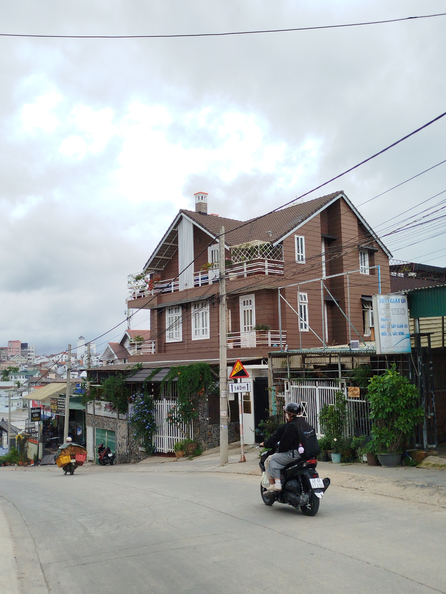 The image depicts a street scene in Đà Lạt, Lâm Đồng, Vietnam. A three-story brown wooden building stands prominently on the corner of the street with white framed windows and balconies with white railings and flower boxes. A street sign indicating a 140-meter distance and a triangular road sign are posted near the building. A scooter with a rider wearing a helmet and backpack is in the foreground of the road. A second scooter is farther down the street carrying boxes. Utility poles with overhead wires crisscross the sky. Buildings of various heights and colors are visible in the background. The sky is overcast with a gray, cloudy sky. Various vegetation is visible around the building and along the street. There is a sign in Vietnamese on the right of the frame, which translates to "Cô Tho Laundry".